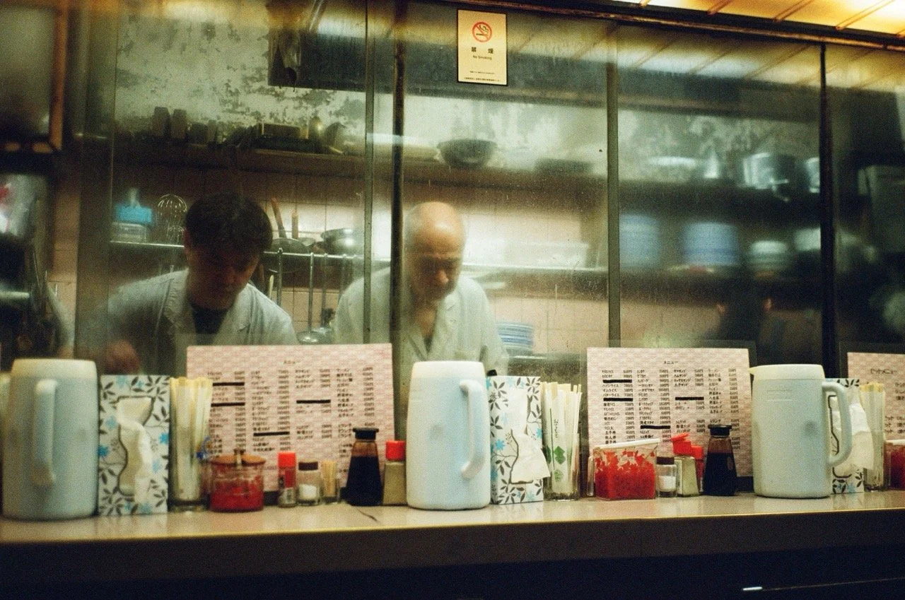 Two chefs working behind a kitchen glass partition, with condiments, napkins, and menus on the counter in front of them.