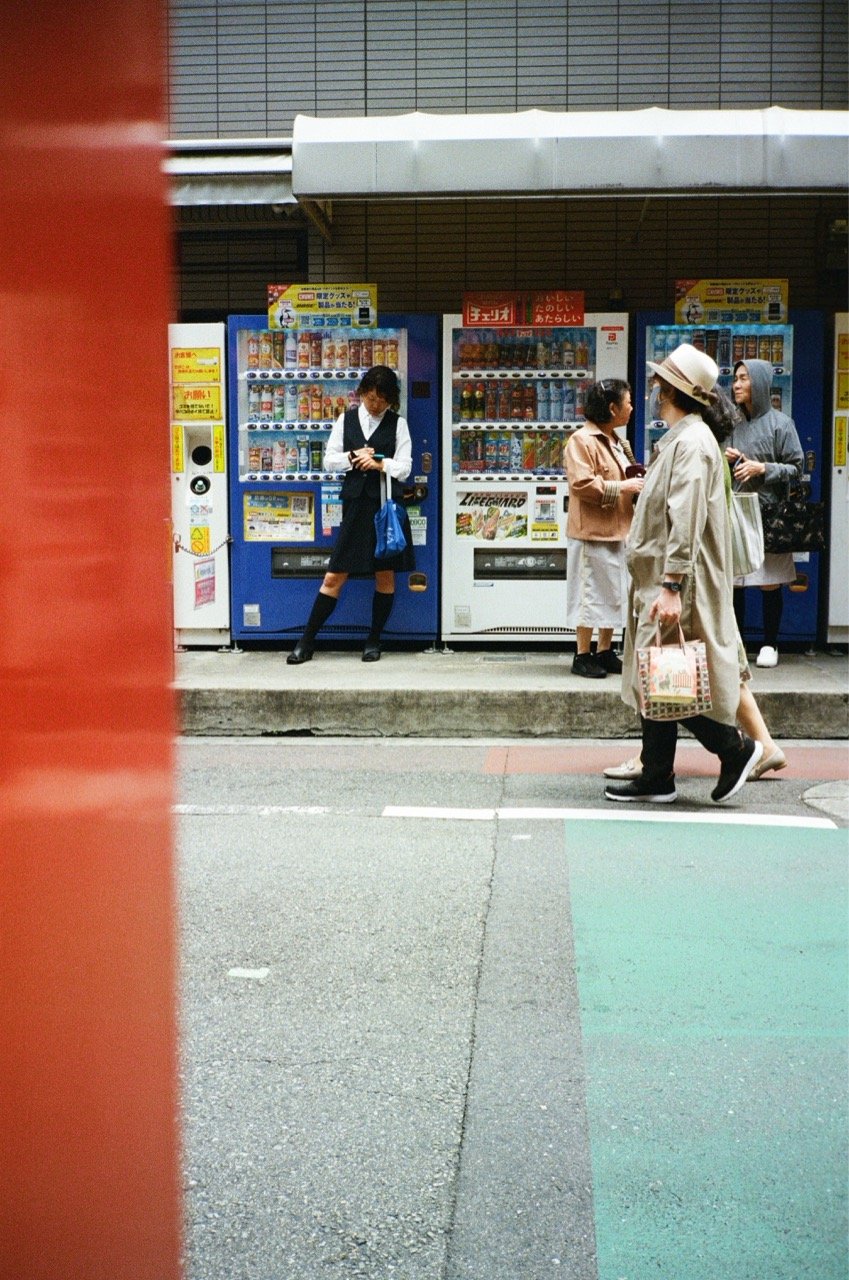 People standing and walking in front of vending machines on a sidewalk, with one person in school uniform and others in casual clothing, some carrying shopping bags.