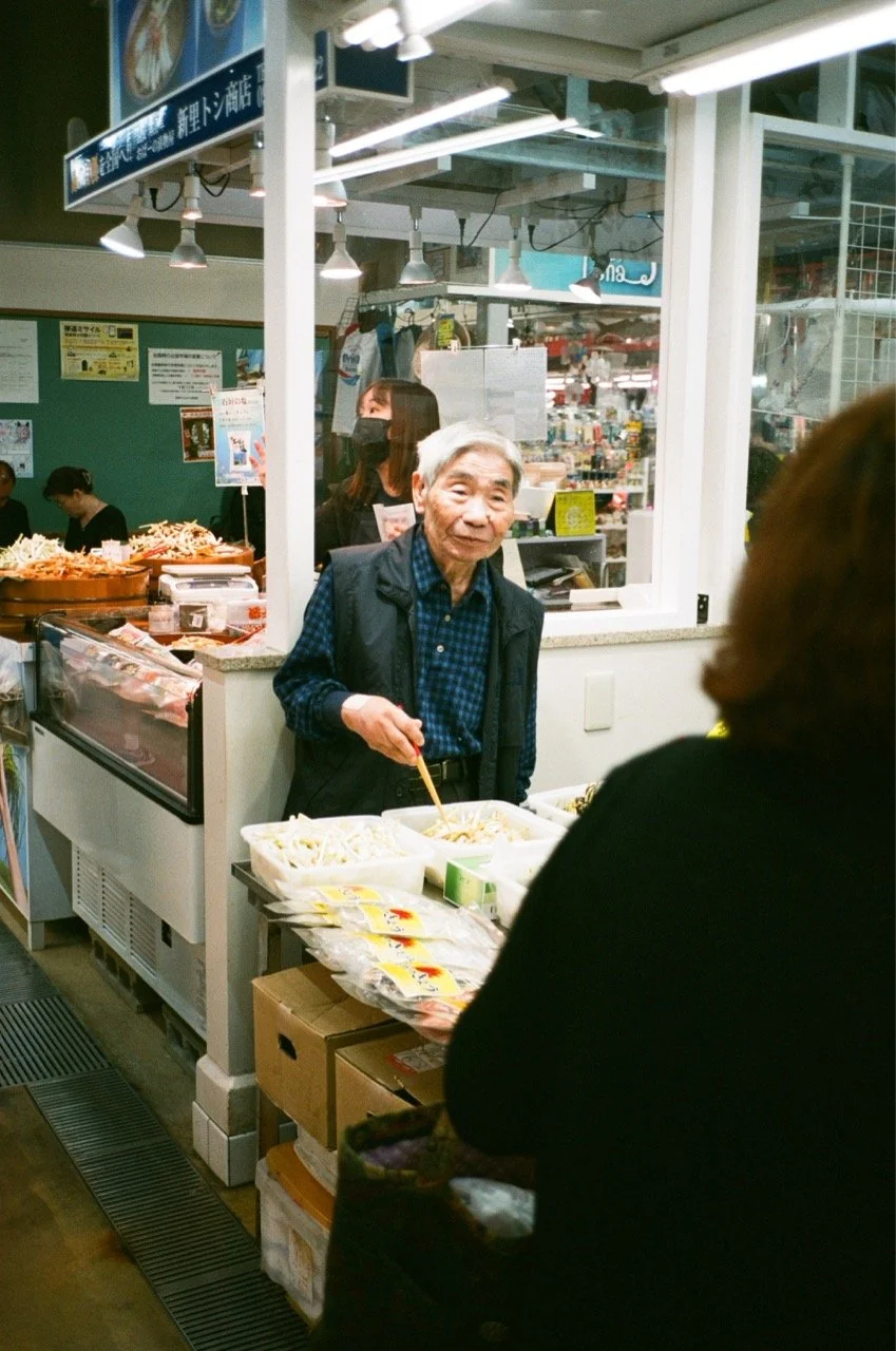 An elderly man selling food at a market stall, with a woman customer in front and a girl driving in the background. The stall has various packaged foods and produce on display.