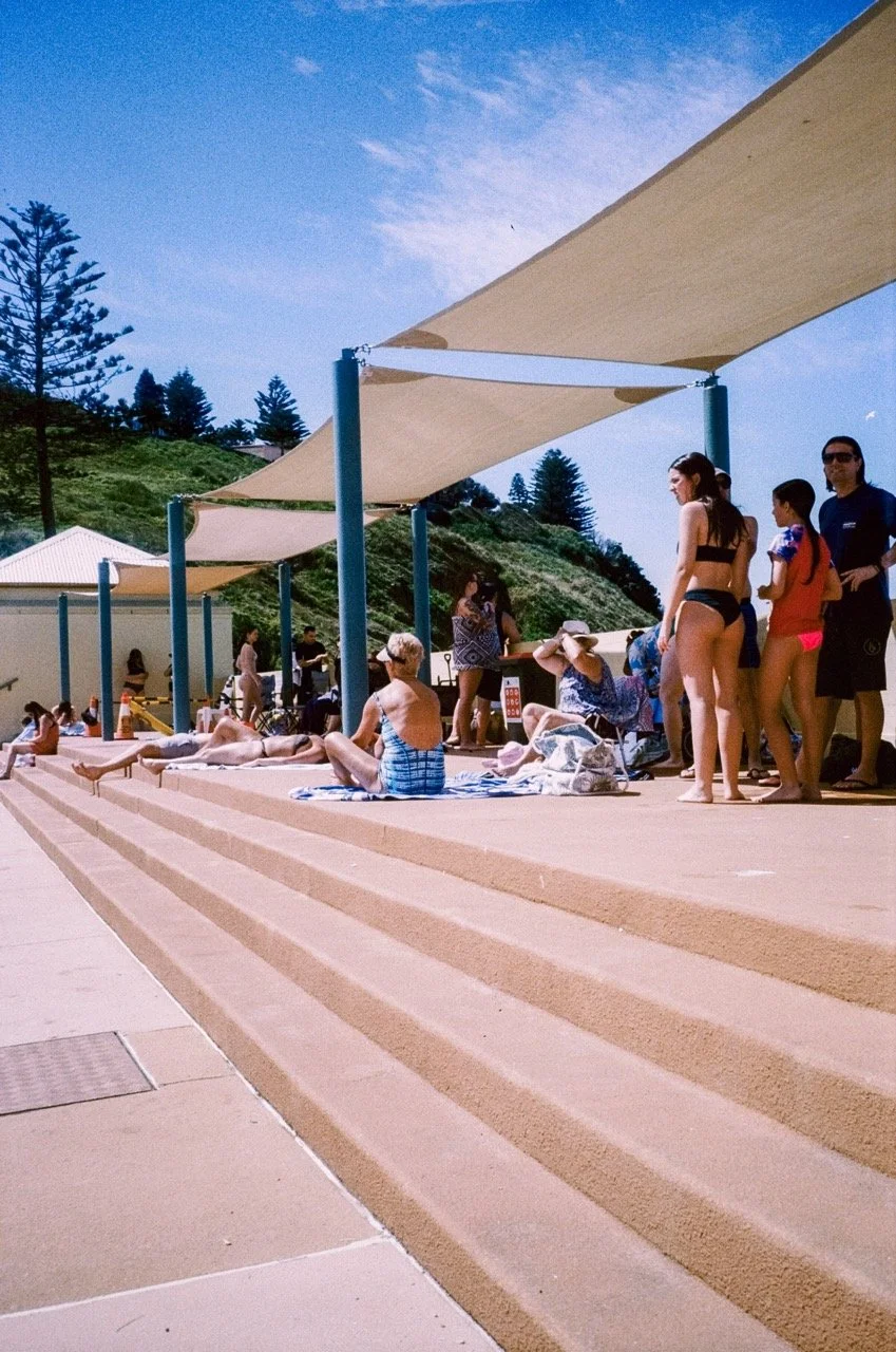 People relaxing and sunbathing at an outdoor pool with shade sails, surrounded by green hills and trees, on a sunny day.
