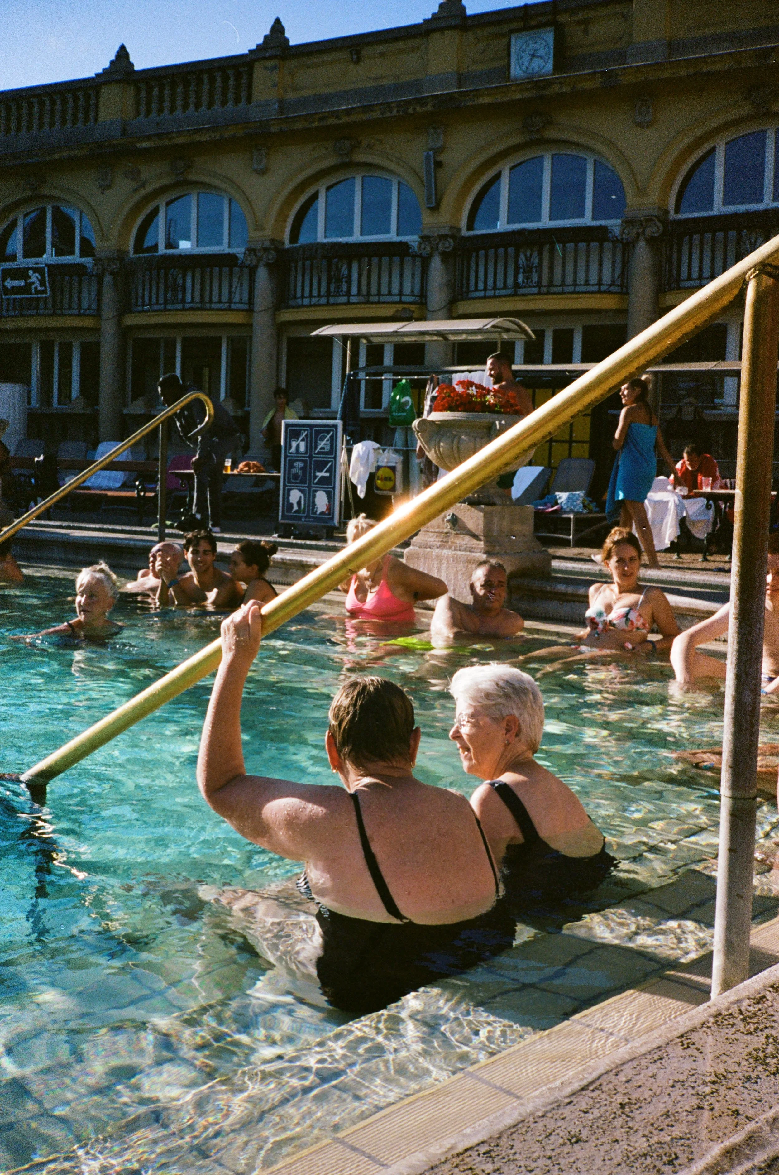 People swimming and relaxing in a communal pool outside a yellow building with arched windows, balconies, and a clock on the wall.