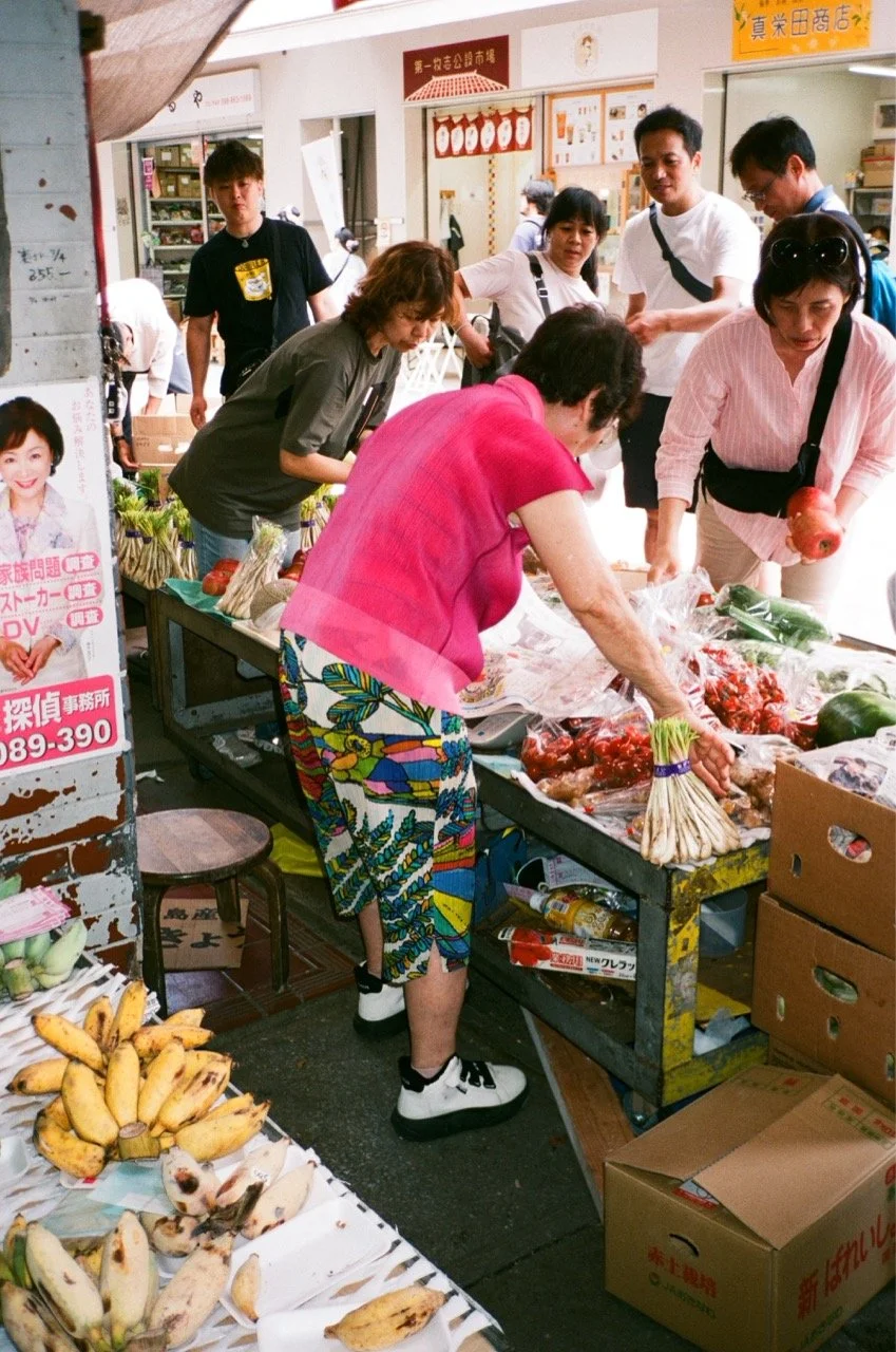 People shopping for vegetables and fruits at an outdoor market stall, with bananas and other produce displayed on tables.
