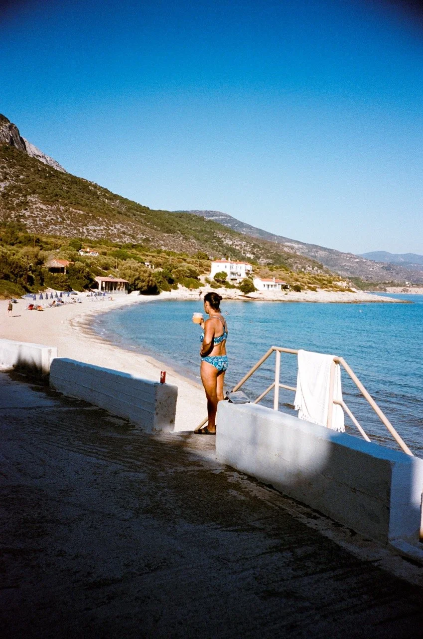 Woman in a blue swimsuit standing on a beach walkway, holding a drink, with a view of a sandy beach, lounge chairs, and green hills in the background.