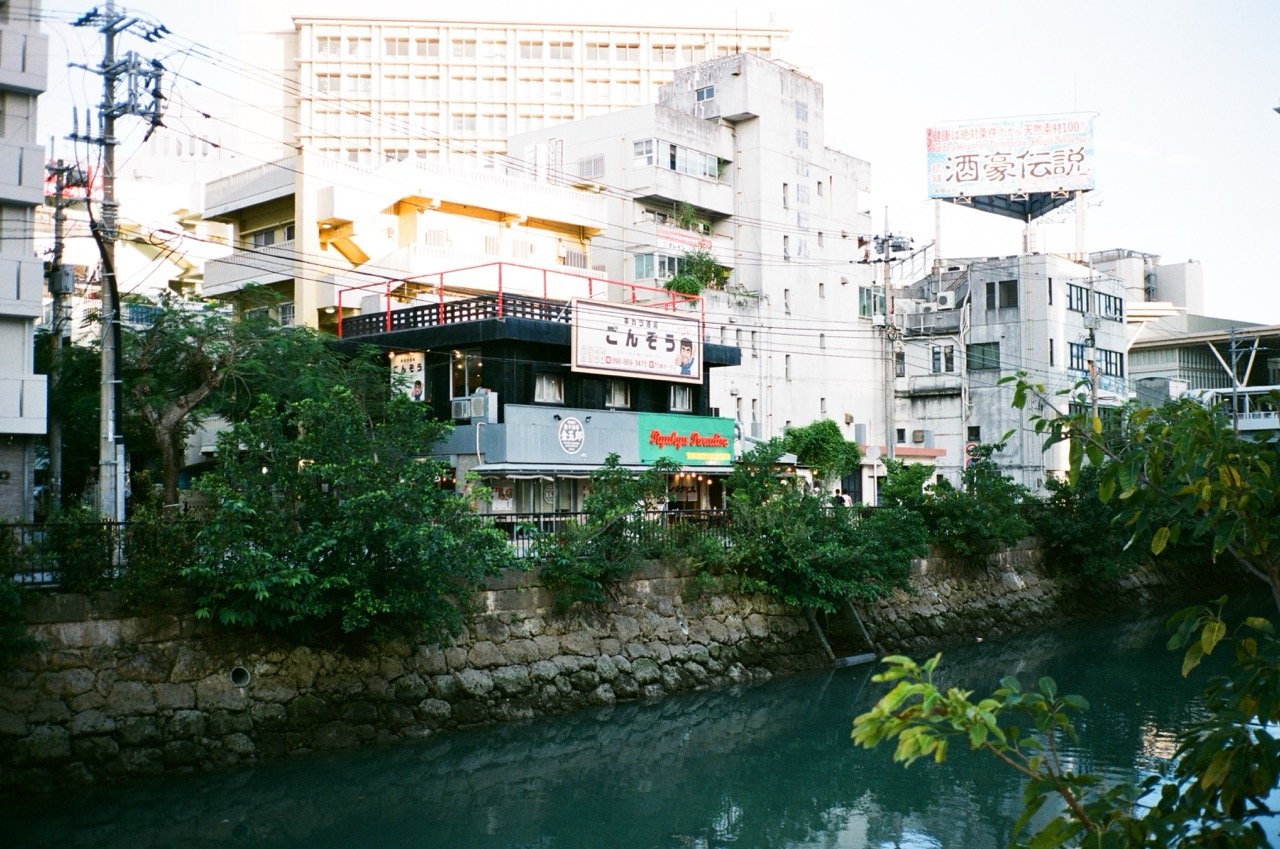 A cityscape with various buildings and greenery along a river, featuring street signs and utility poles.