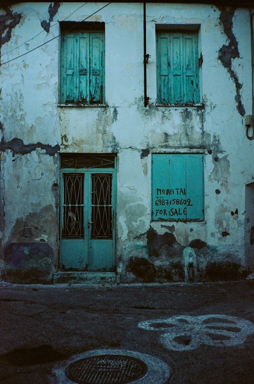 A weathered building with faded turquoise wooden shutters on the windows, peeling paint, and a for sale sign with a phone number written in black, located behind a metal gate on the door.