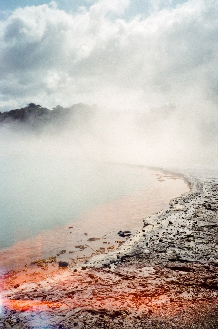 A foggy shoreline with rocky and sandy terrain, mist over water, and cloudy sky.