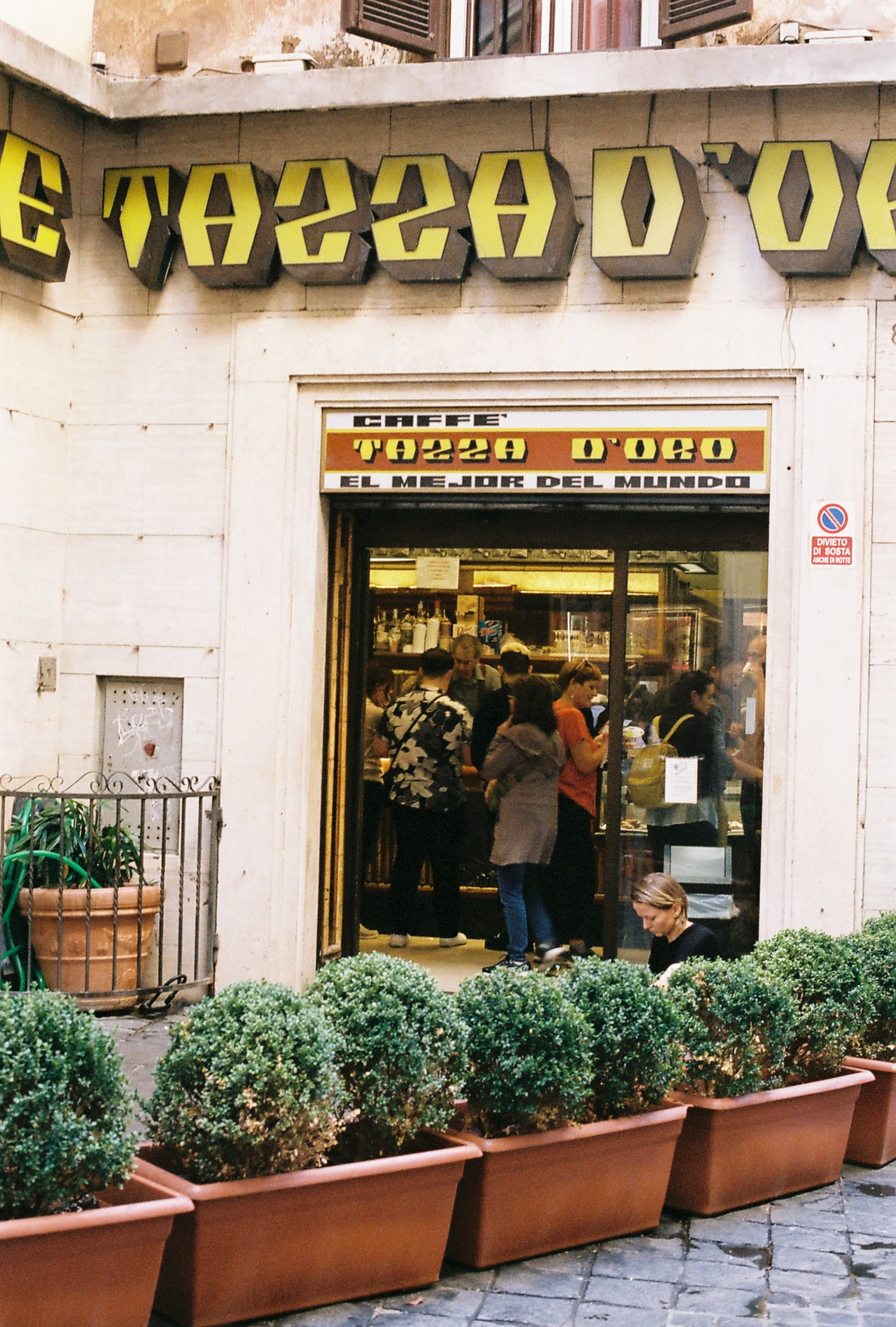 People inside a small shop named 'Tazza D'Oro' with potted bushes outside, selling coffee, with a sign indicating 'The best in the world'.