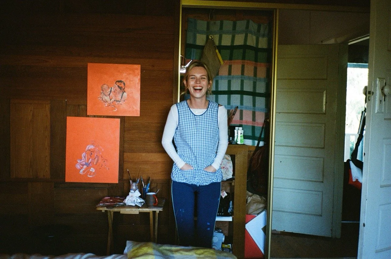 A young woman laughing and standing with her hands in her pockets inside a room with wooden walls, colorful artwork hanging on the wall, and an open closet door beside her.