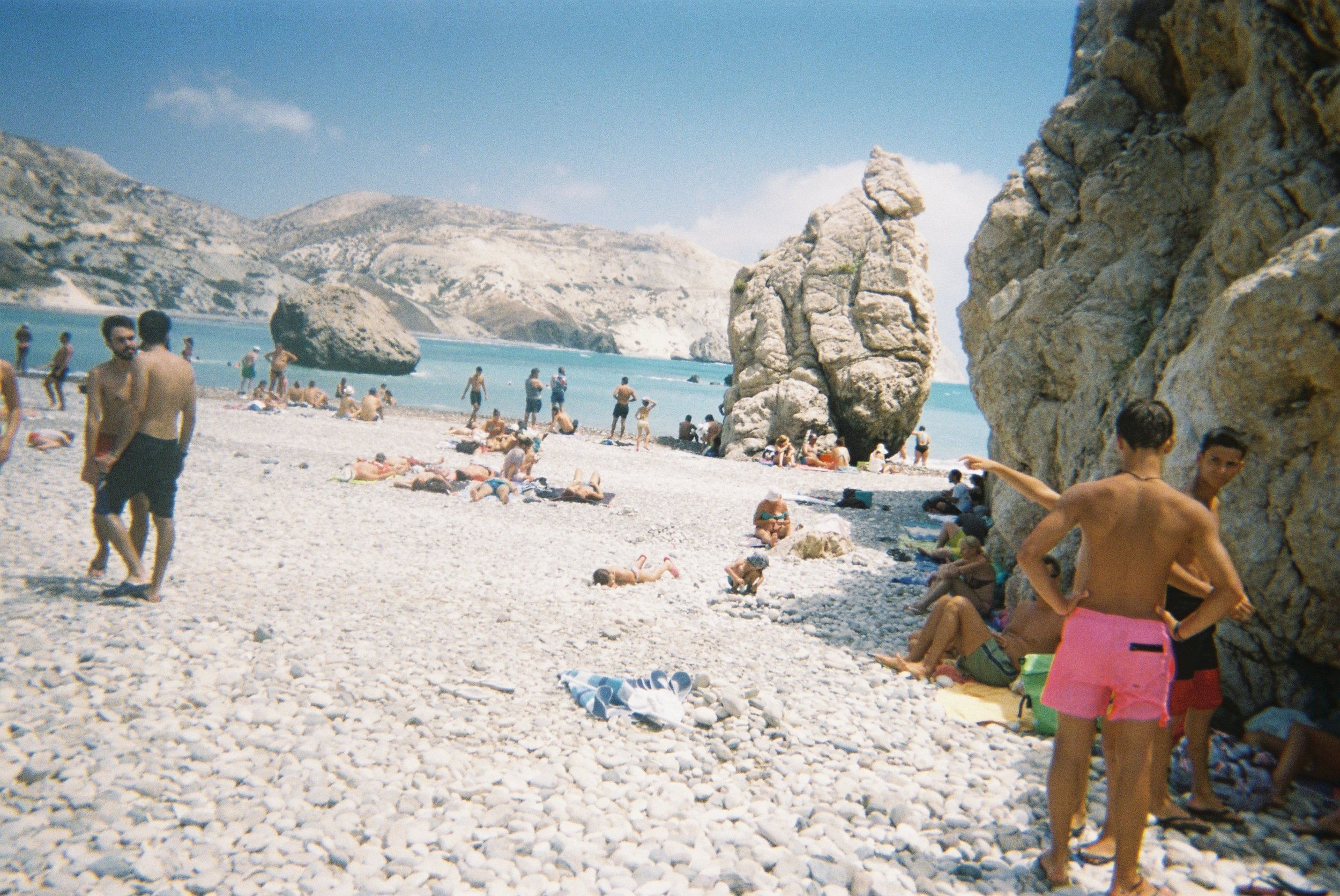 People relaxing and sunbathing on a pebbled beach with large rocks and mountains in the background under a partly cloudy sky.