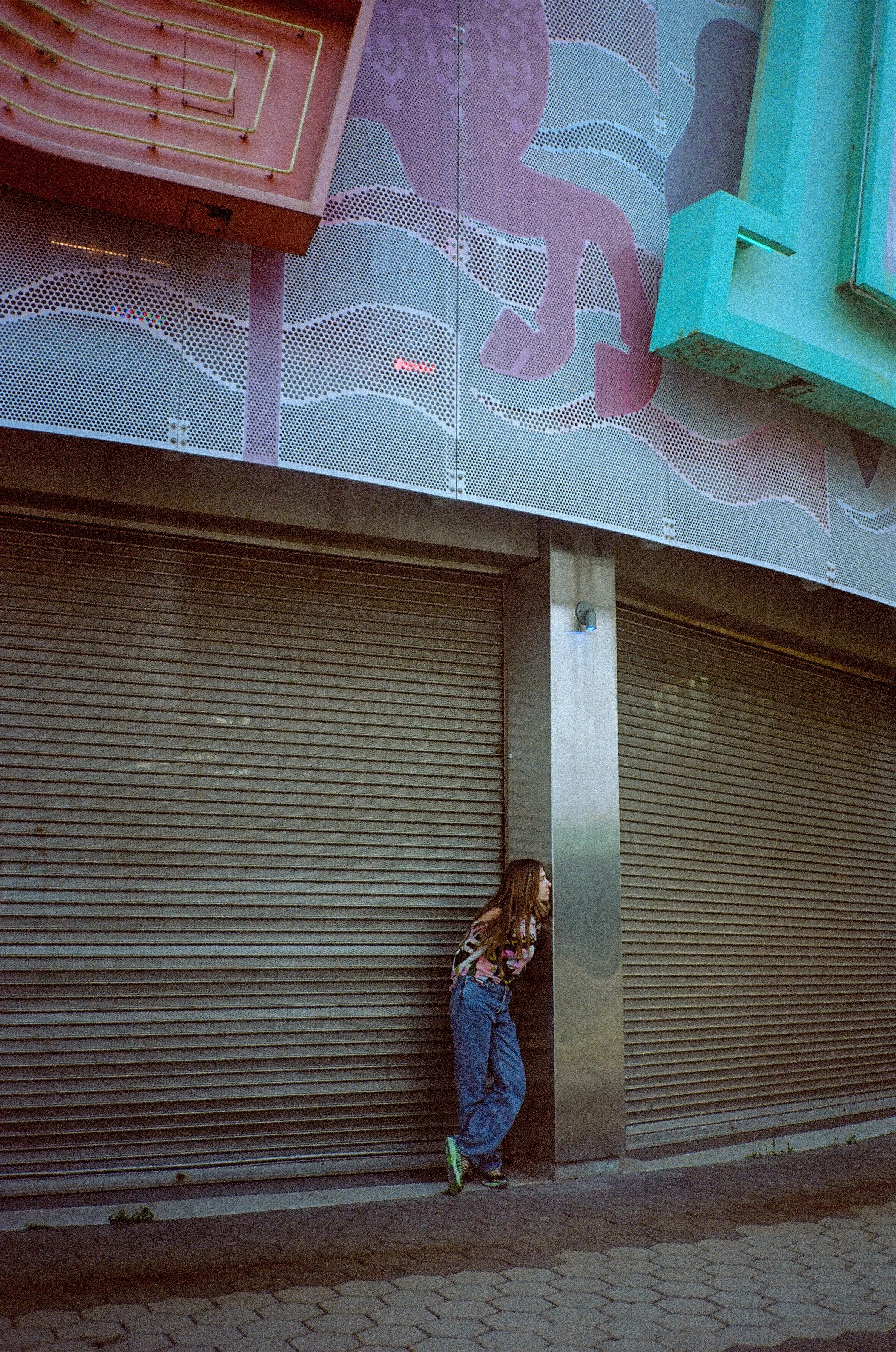A woman leaning against a metal column outside a closed store with a roller shutter door, with a colorful graffiti mural on the upper part of the building's facade.