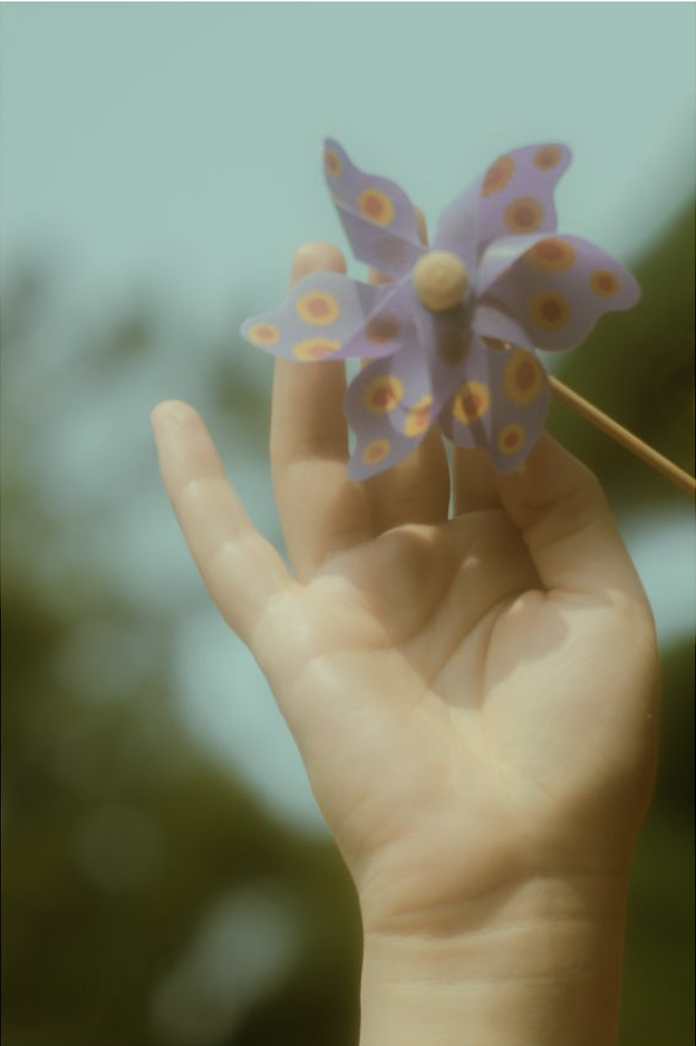 A hand holding a purple paper pinwheel with yellow and orange flower patterns against a blurred green and blue background.