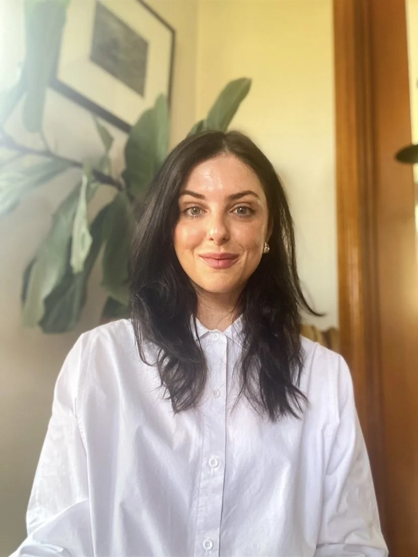 A woman with dark, wavy hair and fair skin smiling at the camera, wearing a white button-up shirt. Behind her, there is a large plant with broad leaves and a framed picture hanging on a wall, all in a warmly lit room.