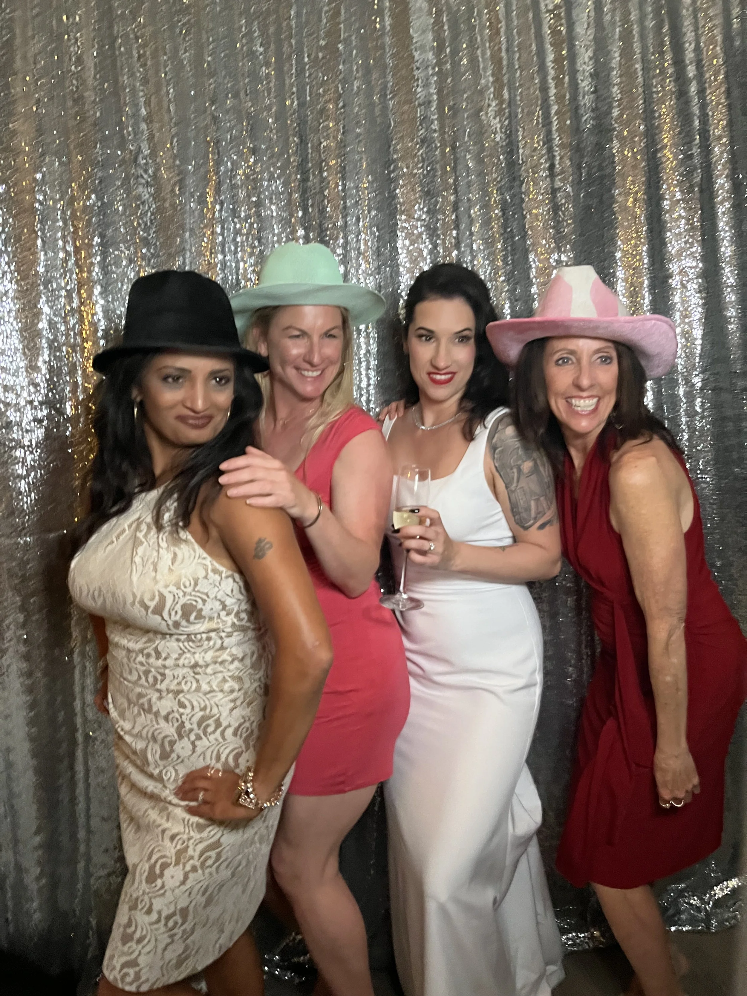 Four women dressed in elegant attire and wearing colorful hats, standing close together with a shiny, sequin-covered backdrop.