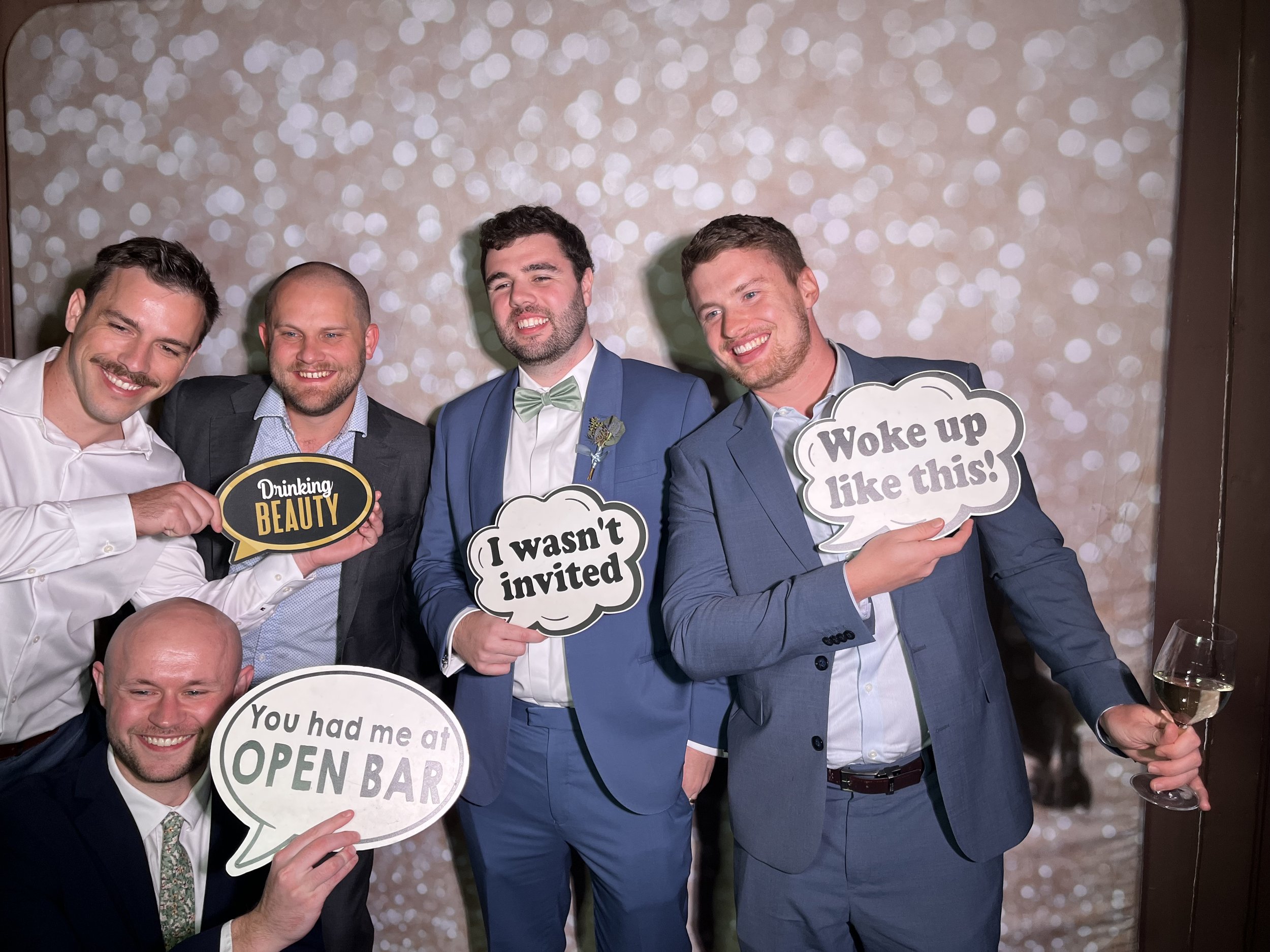 Group of men dressed in formal attire at a celebration or party, holding speech bubbles with humorous phrases, including one that says 'Woke up like this!' and another that says 'Drinking beauty.' The background features a bokeh light pattern.