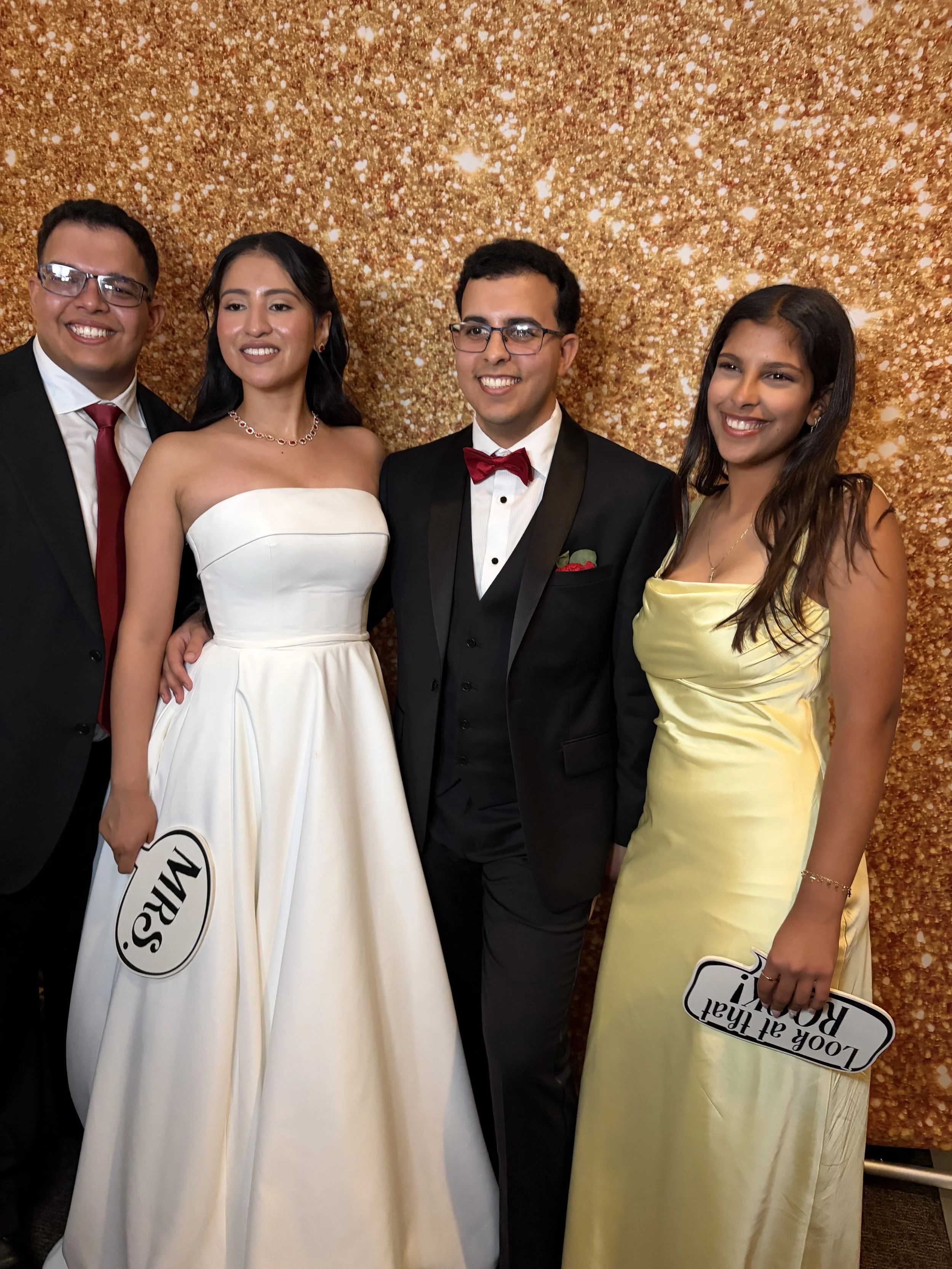 Four people dressed formally, two men in tuxedos and two women in elegant dresses, standing in front of a gold sparkling backdrop at an event.