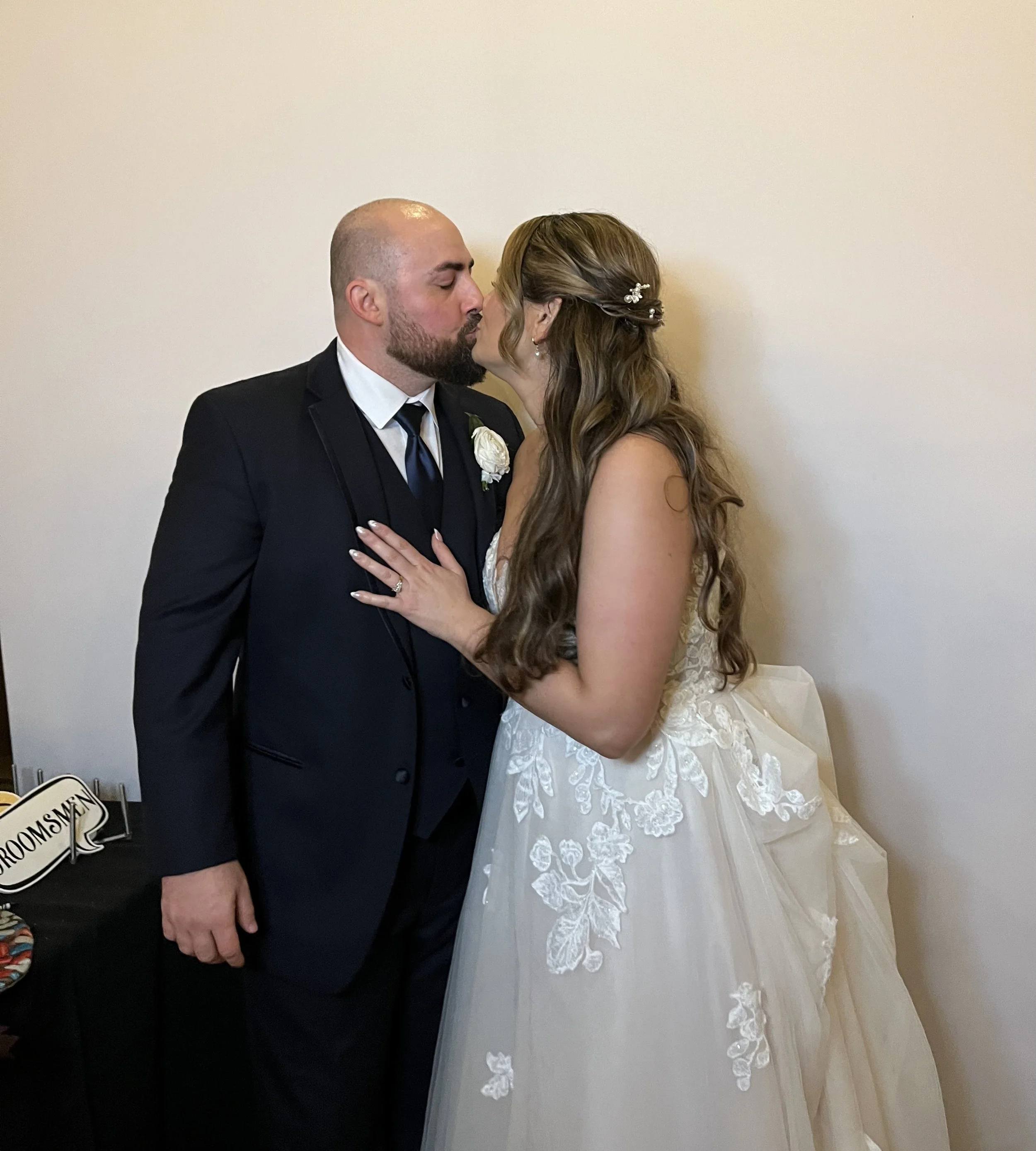 A bride and groom sharing a kiss at their wedding, standing against a plain white wall. The groom is wearing a black tuxedo with a white shirt and a black tie, with a flower boutonniere. The bride has long wavy hair with hair accessories, and is wearing a lace wedding gown. Her hand, with a wedding ring, rests on the groom's chest.