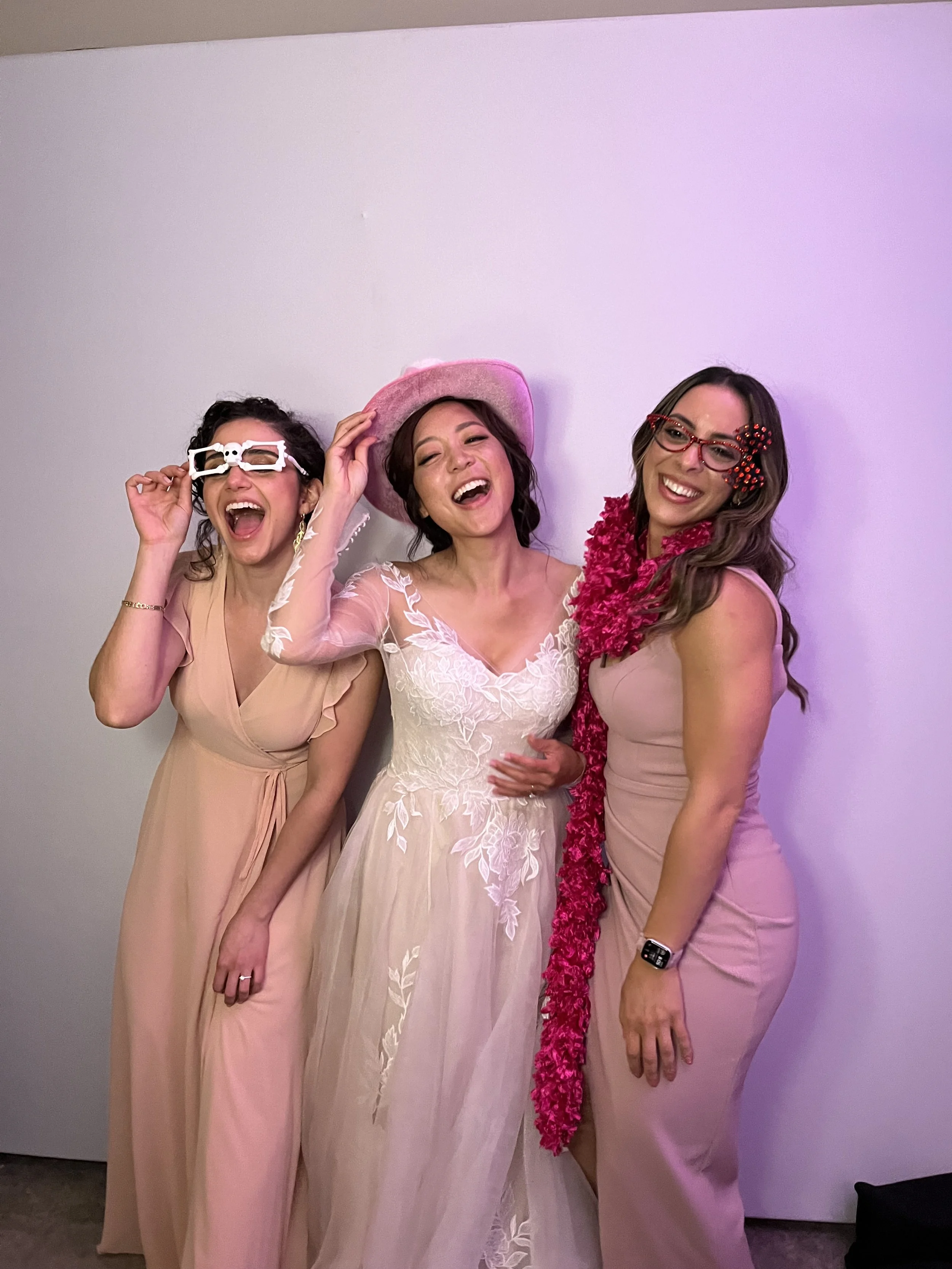 Three women dressed in pink and white at a celebration or party, laughing and smiling. One woman in the center wears a pink hat and a white lace dress. The woman on the right has a pink dress with a red bow and floral accessories. The woman on the left wears glasses with a skull design.