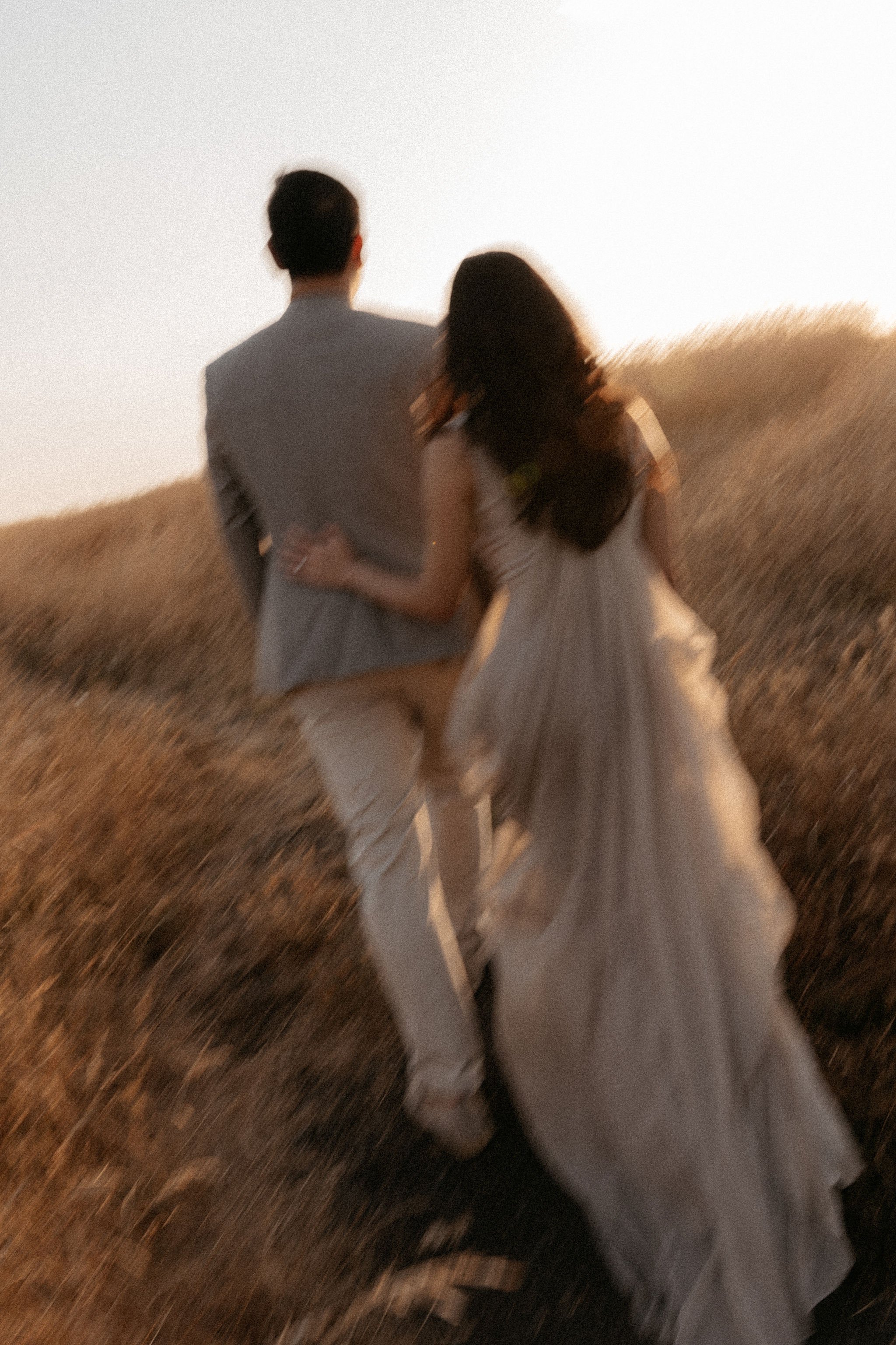 A man and woman dressed in formal attire walking through a field at sunset, with the woman holding the man's hand.