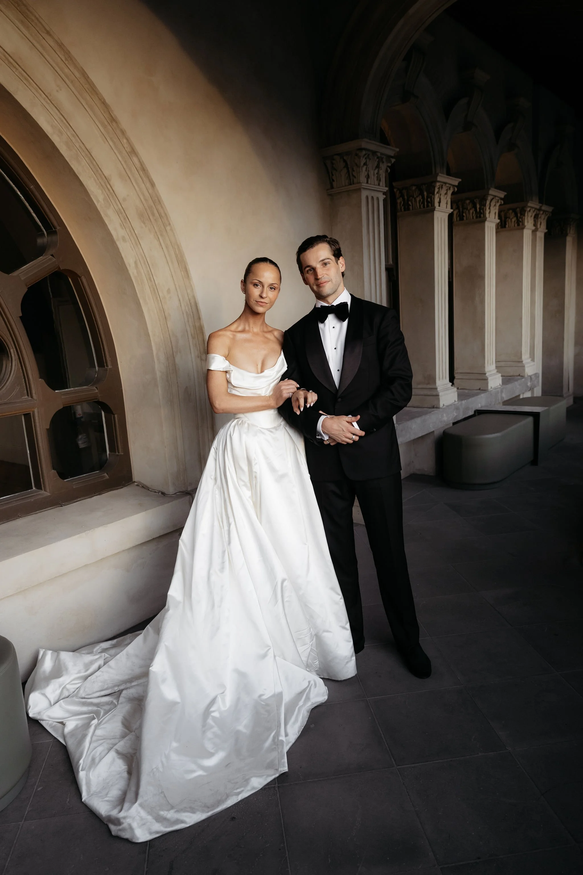 A bride and groom standing inside a church with classical architecture, the bride in a white off-shoulder wedding gown and the groom in a black tuxedo, both looking at the camera.