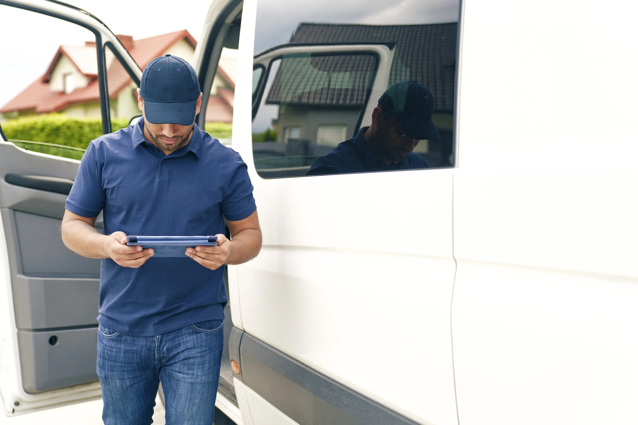 A man wearing a blue cap and polo shirt looking at a tablet next to a white van with an open sliding door.