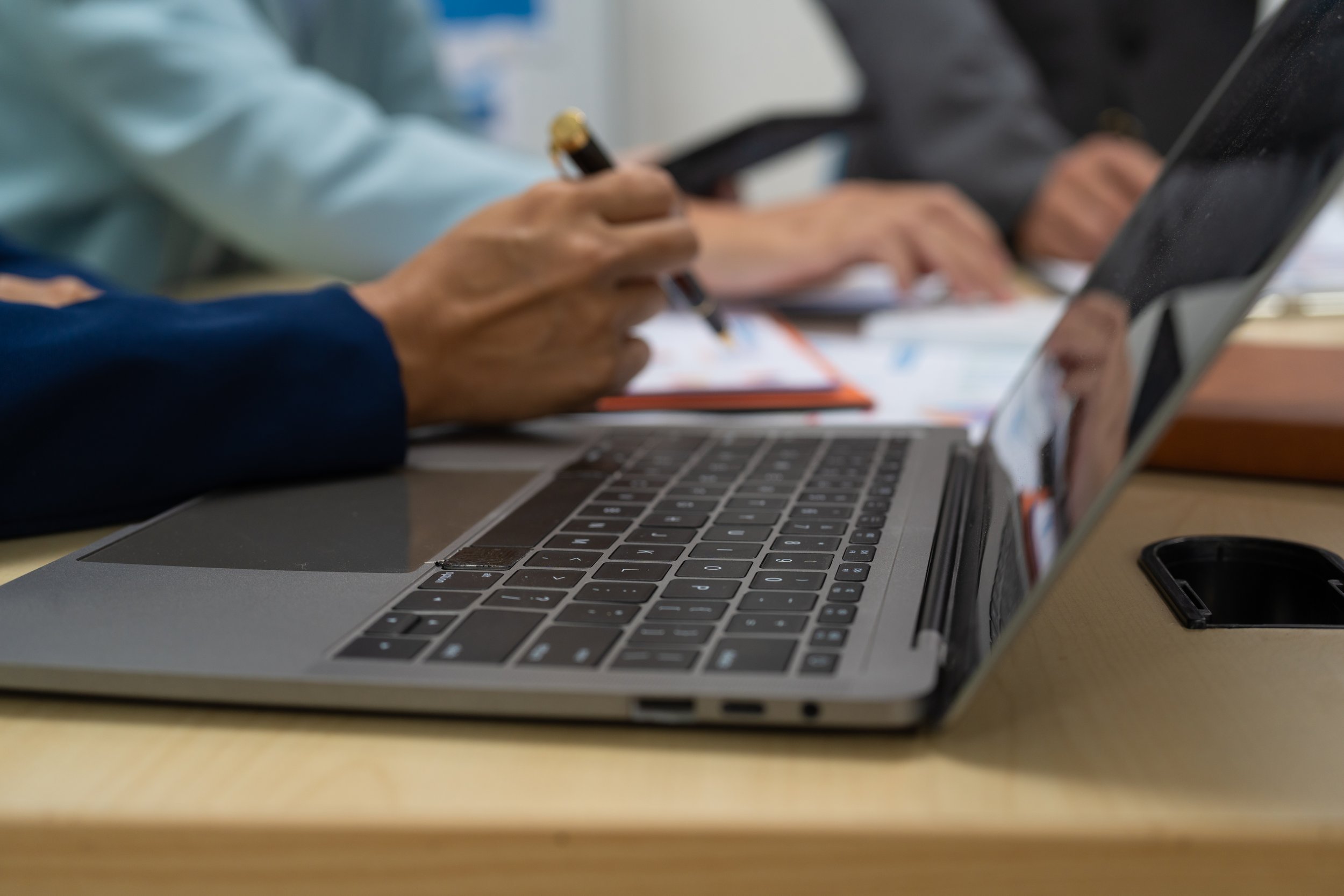 Close-up of a laptop on a desk, with a person's hand holding a pen and taking notes, and another person using a tablet in the background.