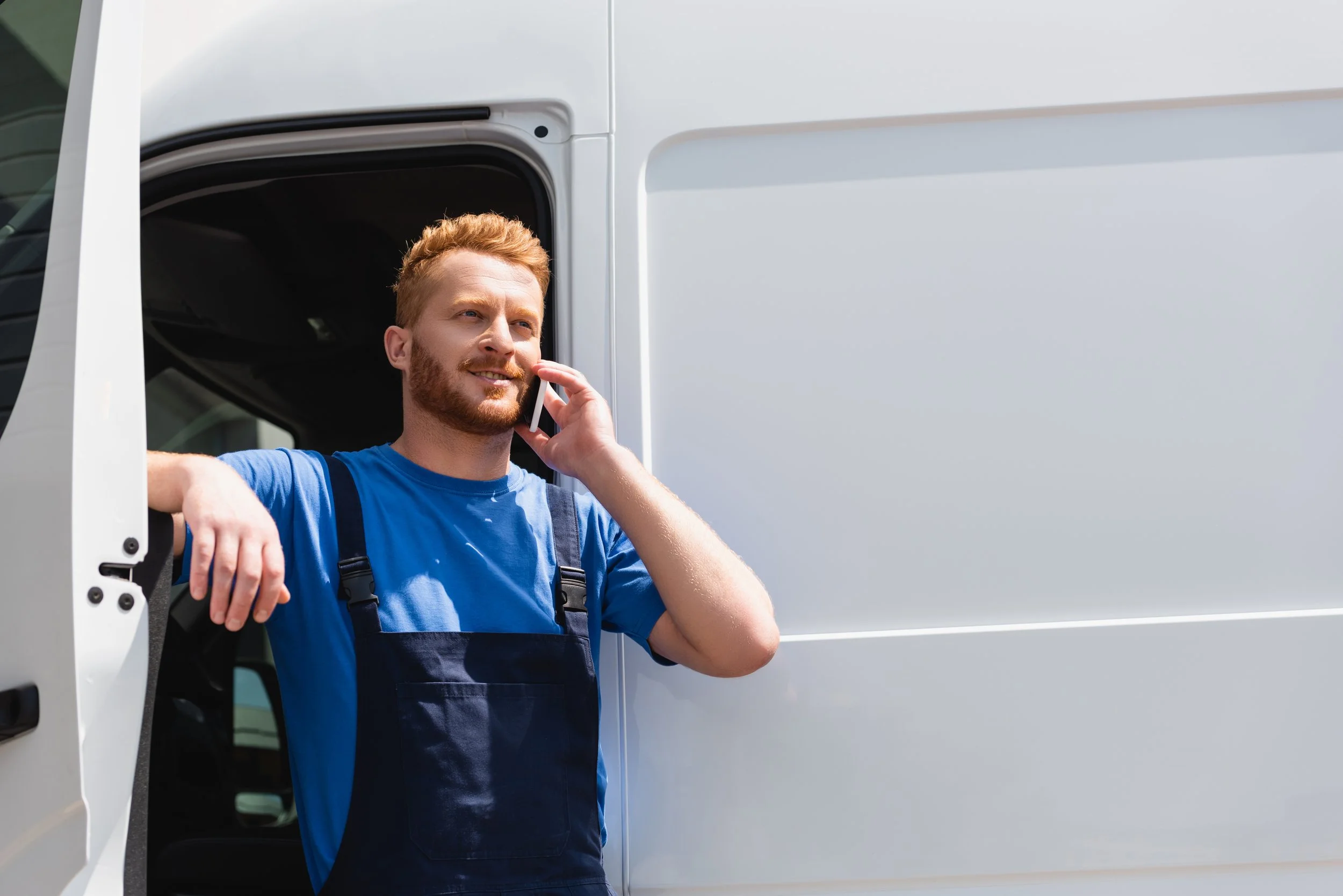 A man with red hair and a beard leaning on the open door of a white delivery truck, talking on his cellphone.