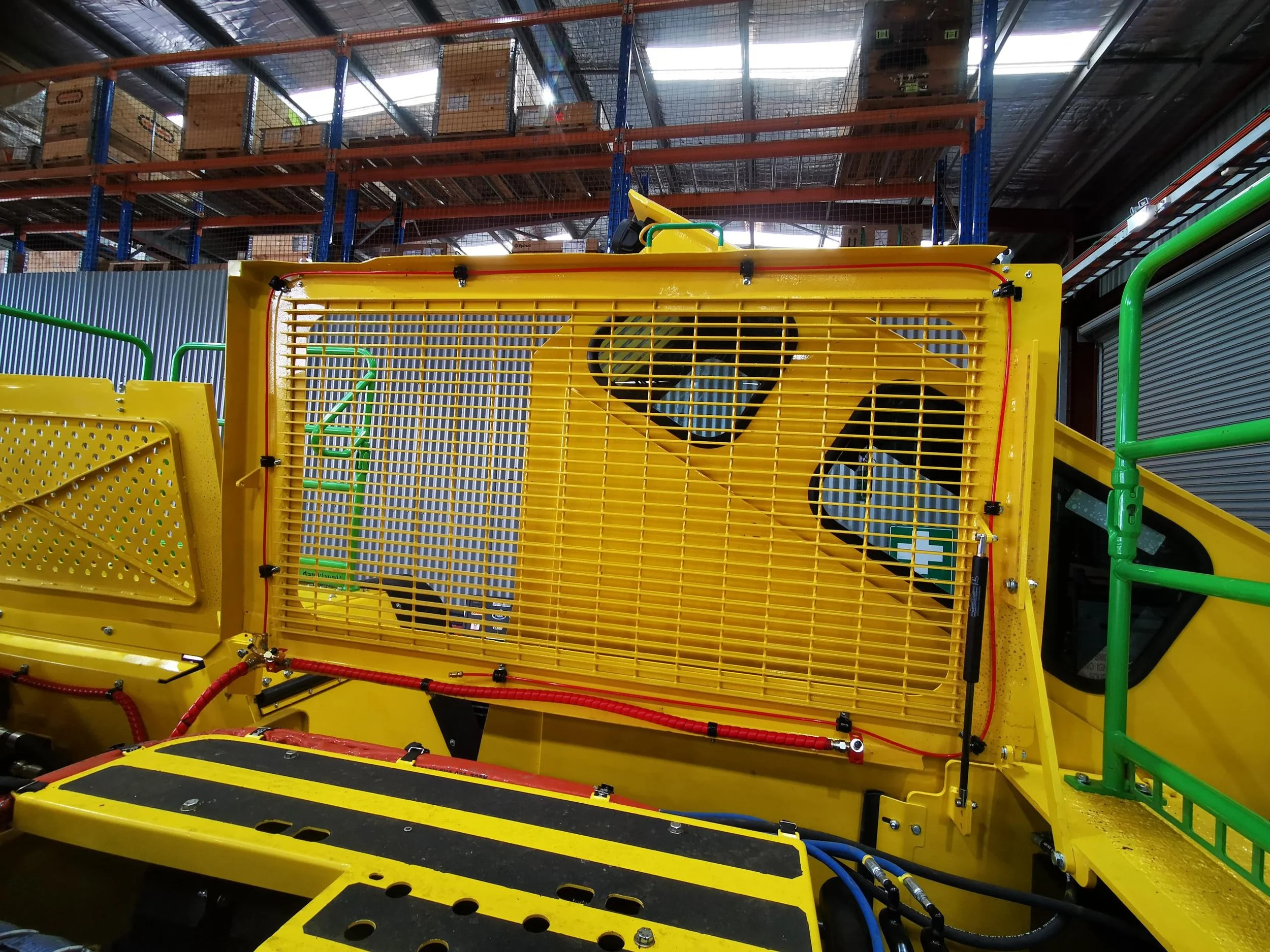 View of a yellow industrial machine with green rails, a grated panel, and various tubes and wires inside a warehouse with shelves and boxes in the background.