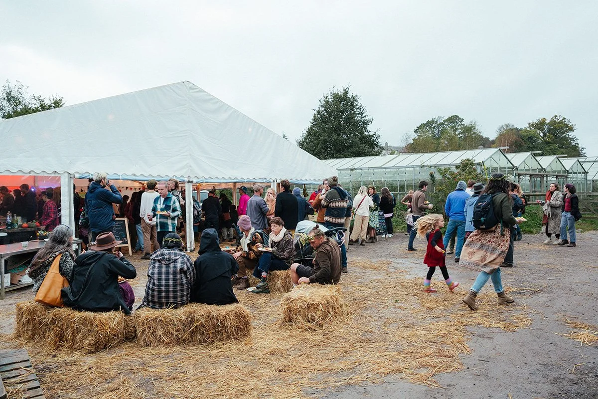 People gathered outdoors at a daytime event with a large white tent and metal greenhouses nearby, some sitting on hay bales, others walking around. The ground is muddy with scattered straw, and there are trees in the background.