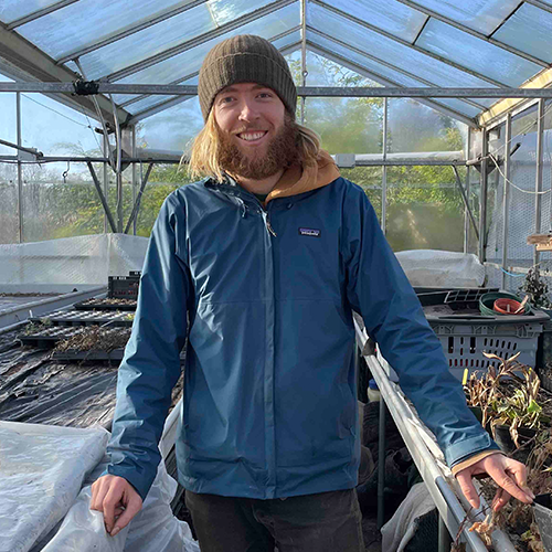 A man with long hair and a beard wearing a brown beanie and a blue jacket standing inside a greenhouse, smiling at the camera.