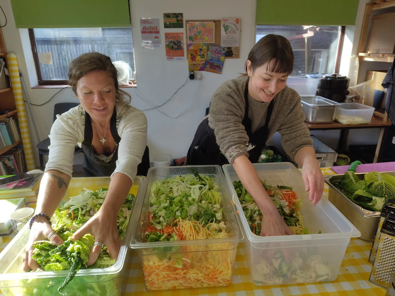 Two women preparing salads with fresh vegetables in a kitchen or a community center, smiling and wearing aprons.