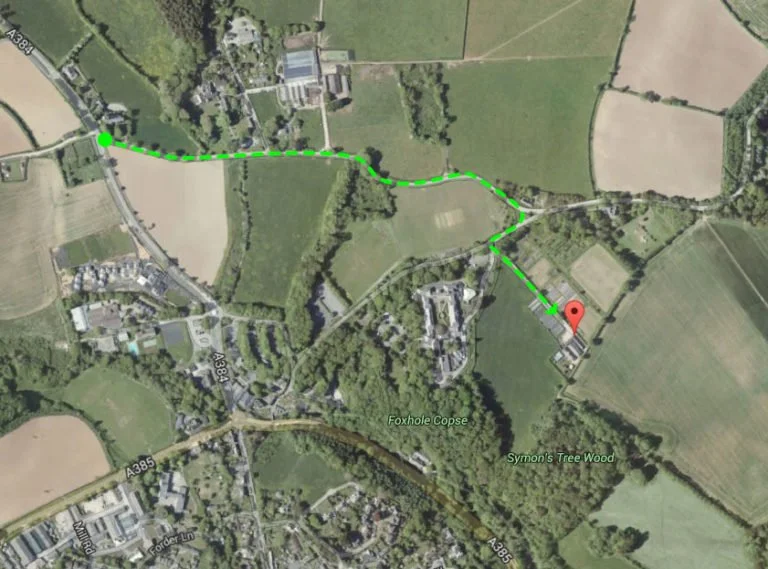 Satellite map showing a green dotted trail path starting from the left side and leading to a red marker on the right, passing through green fields, wooded areas, and residential neighborhoods.
