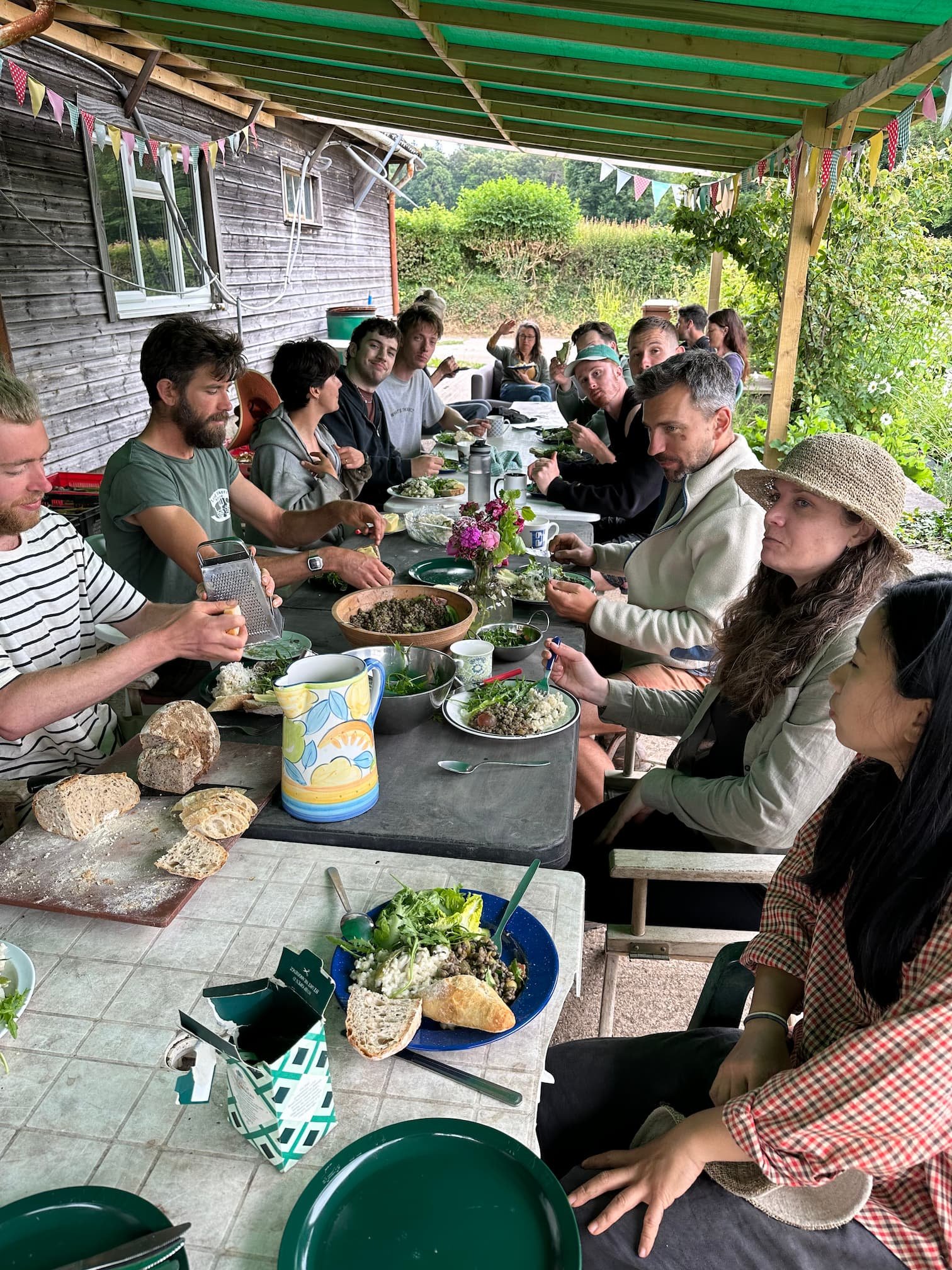 People gathered around a long outdoor dining table under a wooden canopy with colorful bunting, enjoying a meal with bread and salads, in a garden setting.