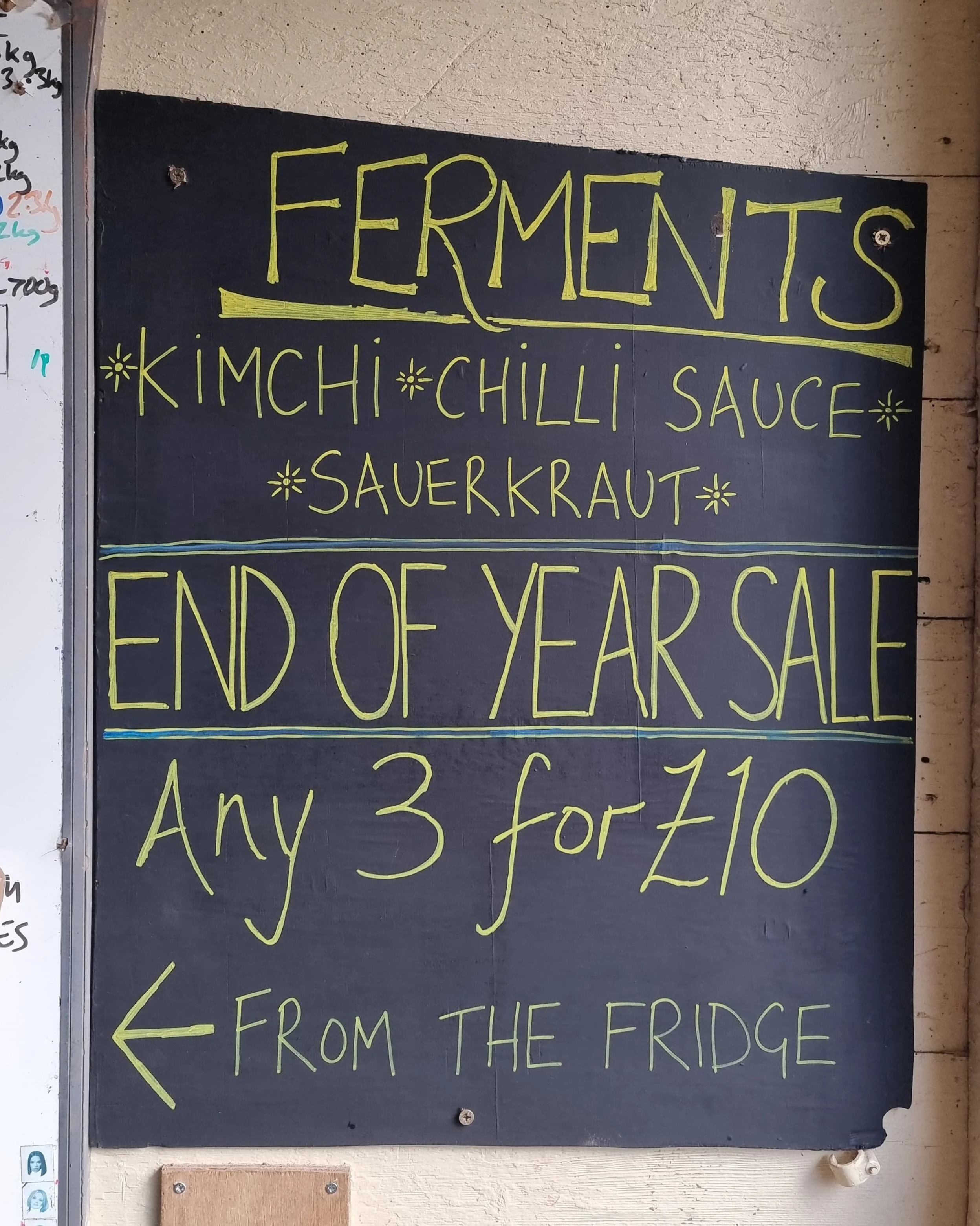 Blackboard sign advertising a year-end sale on ferments like kimchi, chili sauce, and sauerkraut; any 3 for 10 dollars, located in the fridge.
