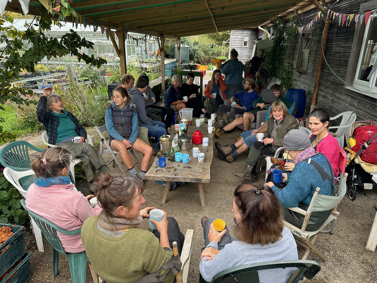 Group of people sitting and socializing in an outdoor garden area under a covered patio, with coffee mugs, teapots, and snacks on a wooden table.