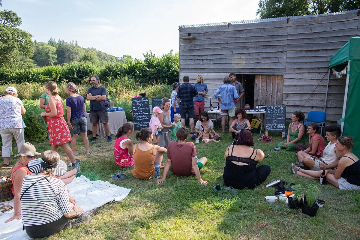 People gathered outdoors near a wooden barn, sitting on the grass and socializing during a summer event with a small outdoor DJ setup.