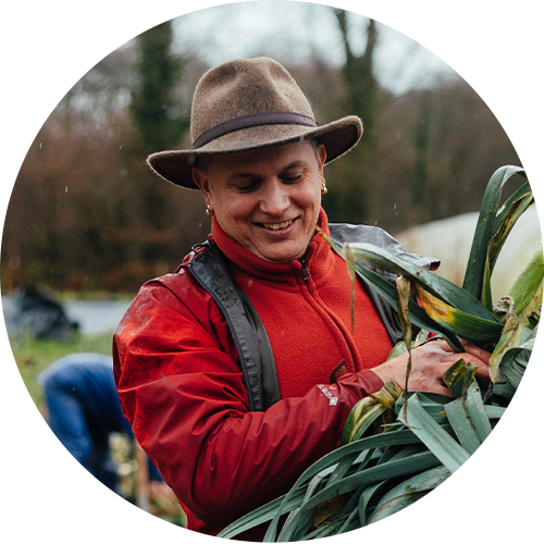 A person smiling while harvesting corn in an outdoor farm setting, wearing a brown hat, red jacket, and black backpack.