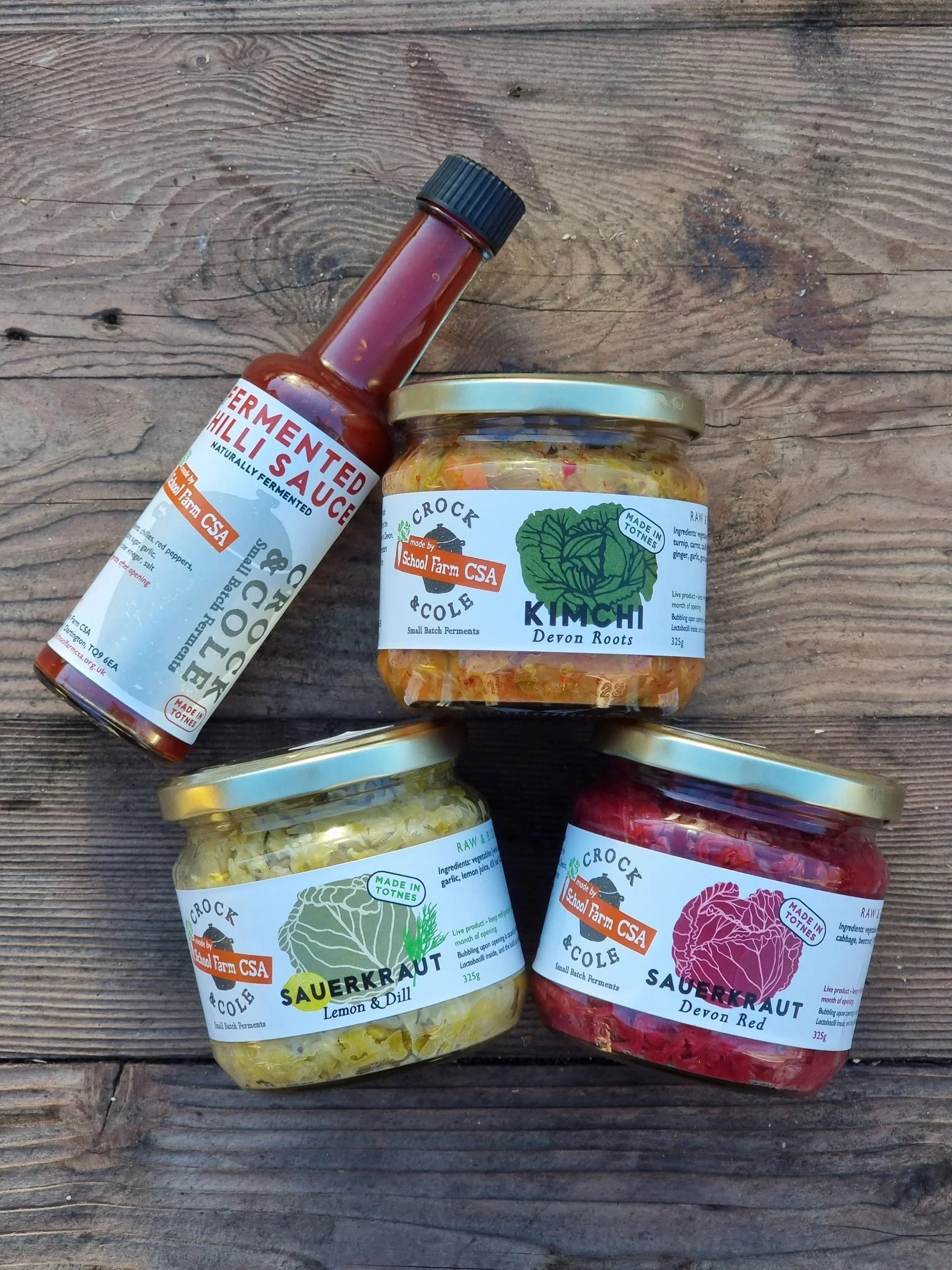 Four jars of homemade fermented vegetables and a bottle of fermented chili sauce on a wooden surface. The jars are labeled as kimchi, sauerkraut, sauerkraut with lemon and dill, and red sauerkraut, all made by School Farm CSA.