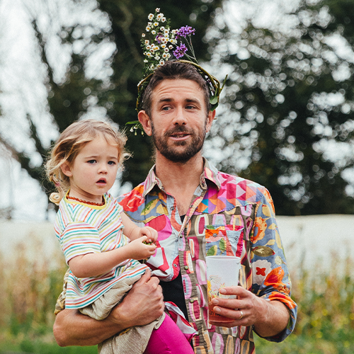 Man with a floral crown holding a young girl in a striped shirt outdoors.