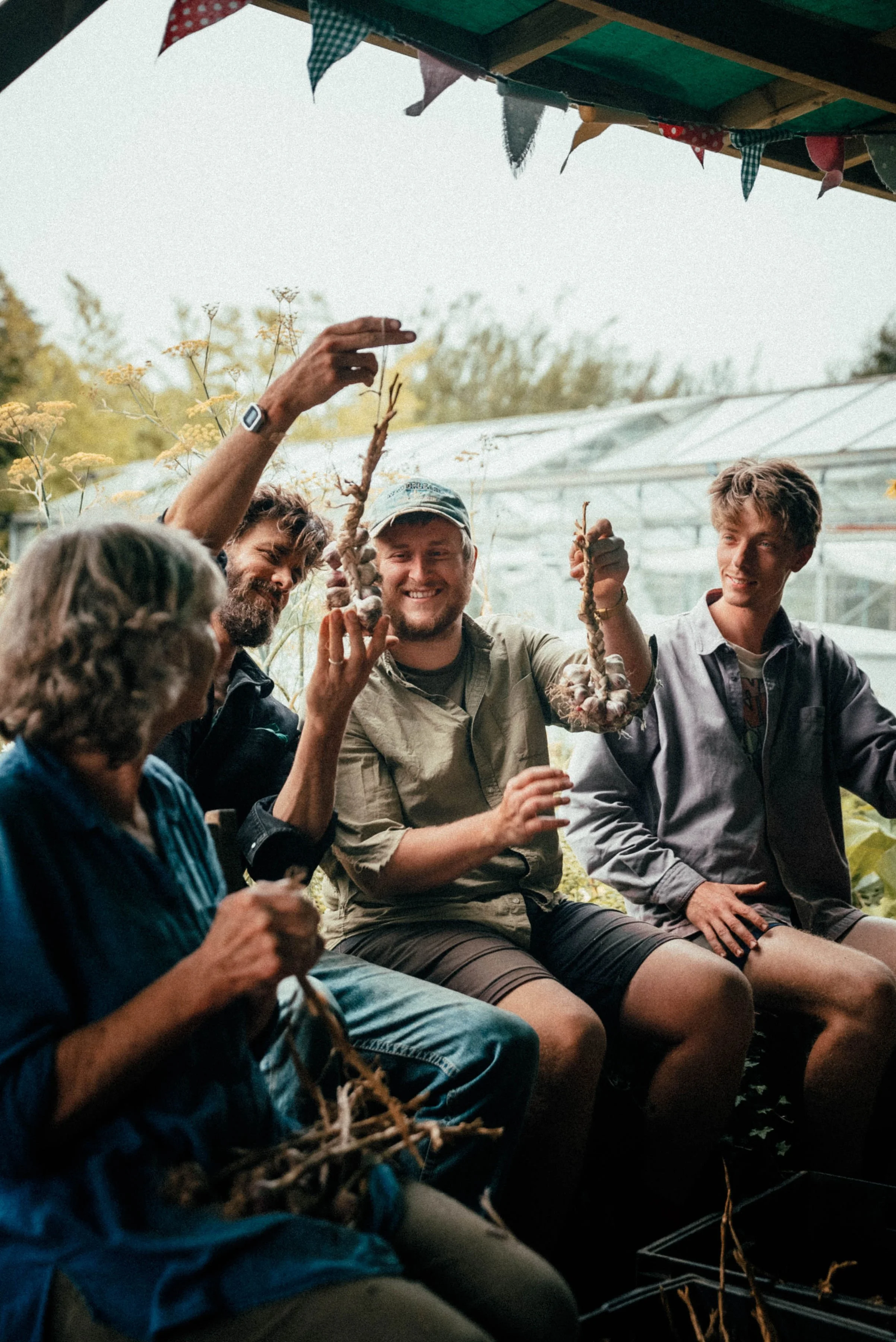 Group of people sitting together, holding freshly harvested garlic bulbs, smiling and enjoying each other's company outdoors under a canopy decorated with colorful bunting.