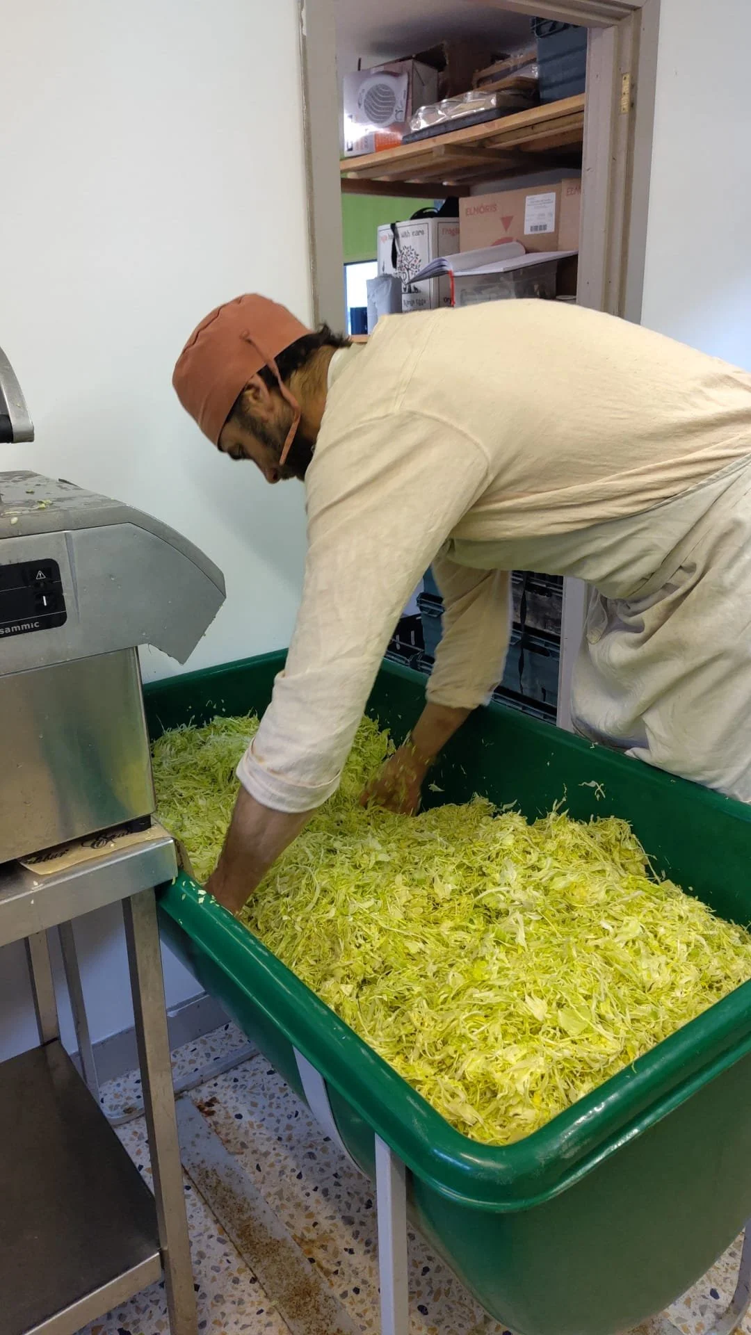 A person wearing a beige long sleeve shirt, white work pants, and a brown head covering is standing in front of a green tub filled with shredded cabbage, working with their hands.