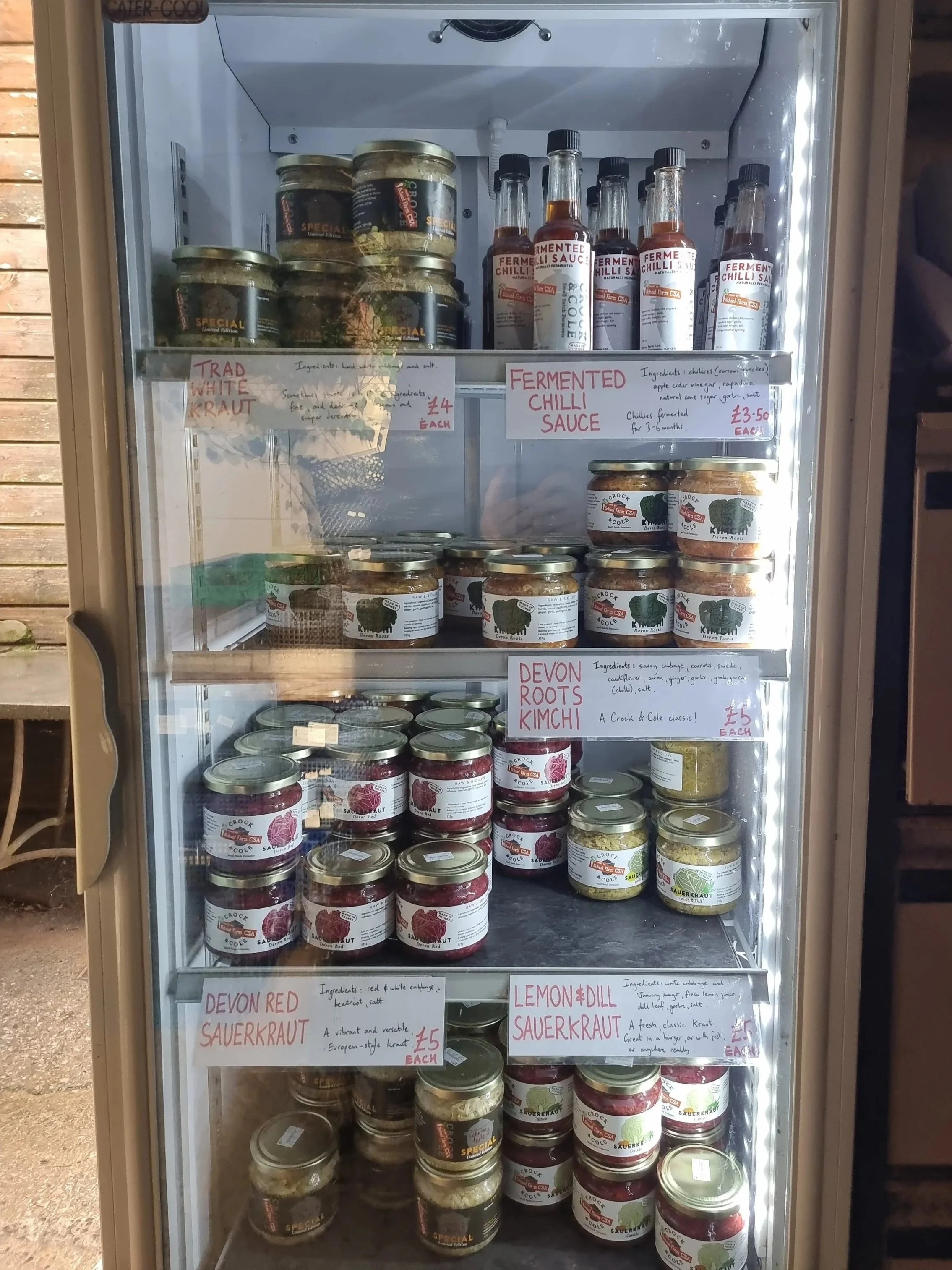 A refrigerated display case with jars of fermented foods including fermented chili sauce, devon roots kimchi, devon red sauerkraut, and lemon & dill sauerkraut, along with bottles of fermented chili sauce on the top shelf. Handwritten signs describe each product and their prices.