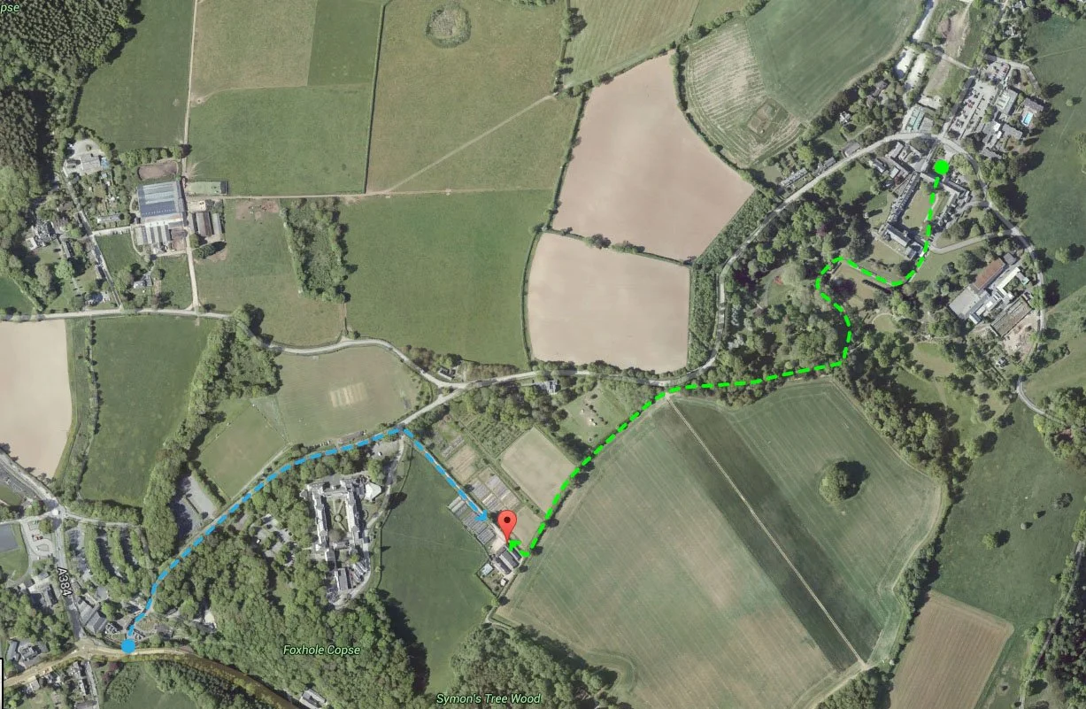 Aerial view of a rural area with fields, trees, roads, and buildings, showing two walking paths starting from the right side of the image, marked with green and blue dashed lines.