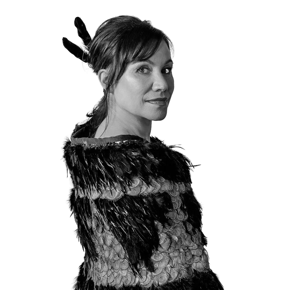 Black and white portrait of a woman in a feathered costume, looking over her shoulder with a confident expression, against a plain background.