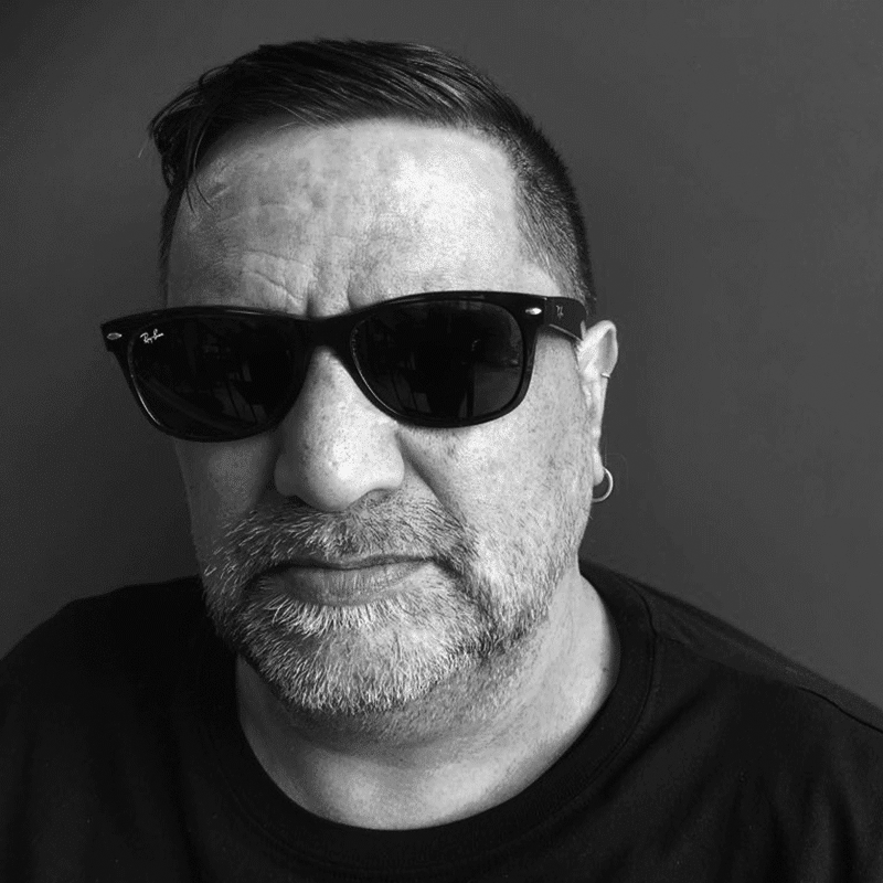 A black and white photo of a man wearing sunglasses and a black shirt, with short dark hair, a beard, and a mustache, looking at the camera.