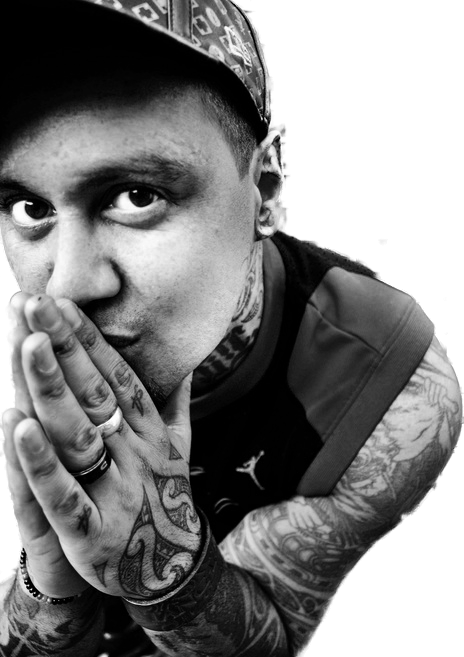 A man with tattoos on his arms and neck, wearing a cap and a sleeveless shirt, holds his hands together near his face and looks directly at the camera in black and white.