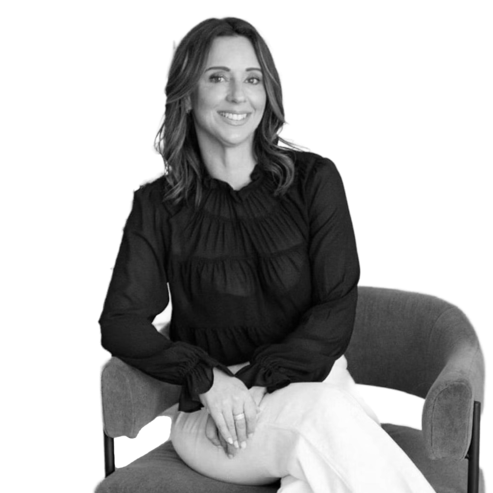 A smiling woman with shoulder-length wavy hair, wearing a black long-sleeve top and light-colored pants, sitting on a cushioned chair with her hands resting on her lap.