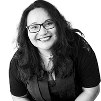 A woman smiling, wearing glasses and a black top, with long, curly hair.