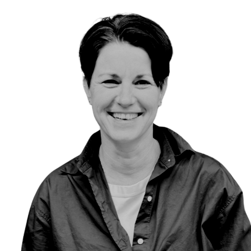 A woman with short dark hair smiling in a black and white photo, wearing a casual button-up shirt over a t-shirt.