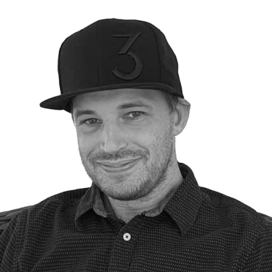 A man wearing a black baseball cap with a number three logo and a dark, collared shirt, smiling at the camera.