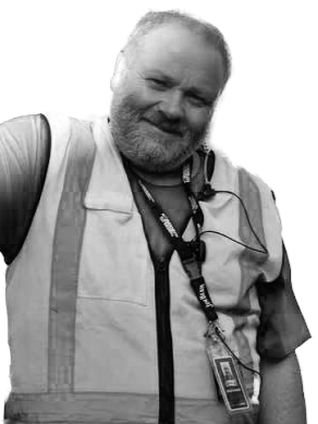 A smiling man with a beard and gray hair, wearing a vest and a lanyard with an ID badge, standing against a plain background.